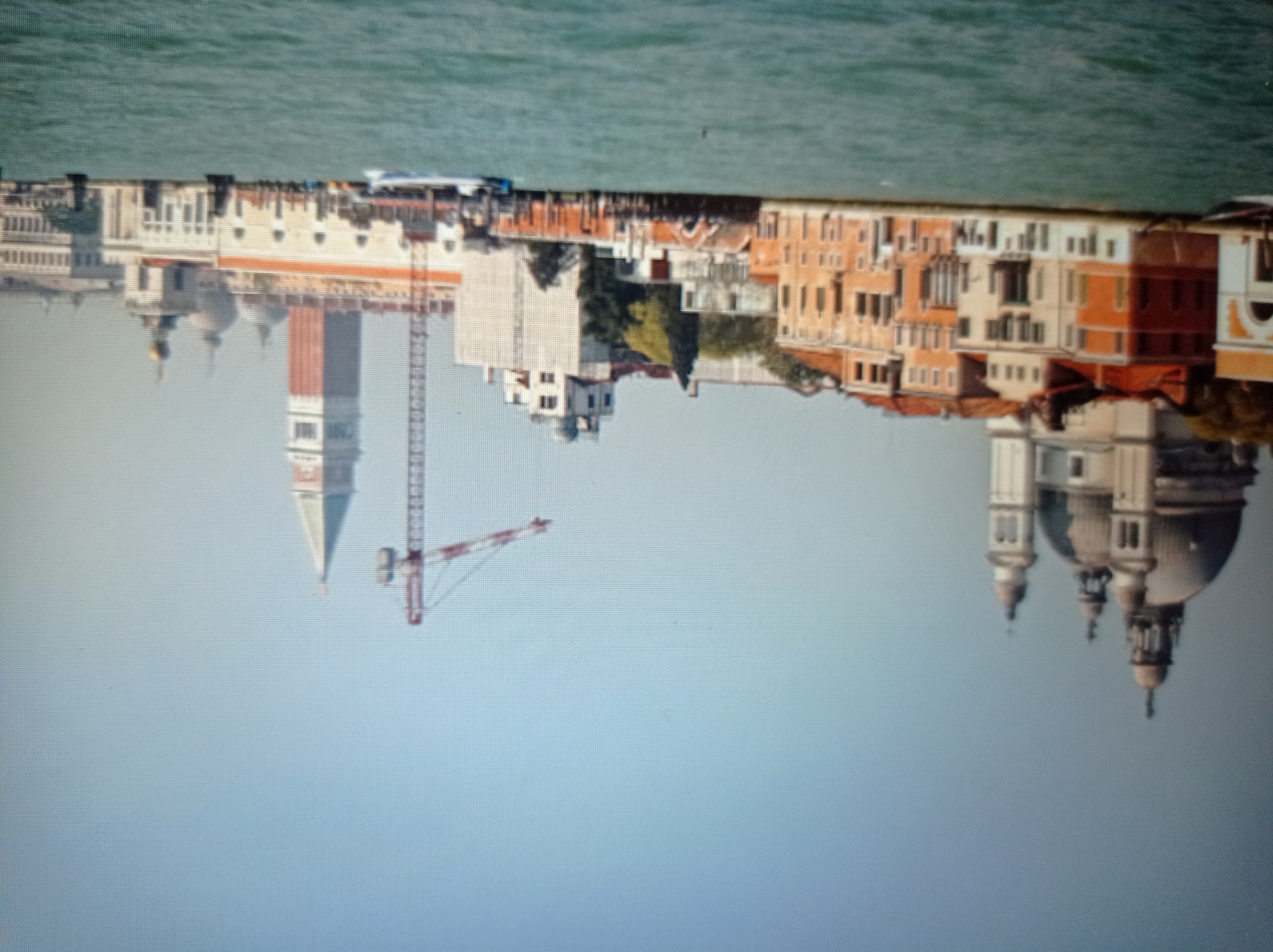 The image shows a wide shot of a waterfront with buildings and a canal in the foreground. On the left, a large domed building with multiple smaller domes and spires is visible. Adjacent to it, a row of multi-story buildings with orange and tan facades line the canal. In the middle ground, a construction crane with red and white accents stands tall against a light blue sky. Further to the right, a slender, red and white bell tower with a green roof pierces the sky, accompanied by more low-rise buildings. The water in the canal is a murky green, with several boats visible, including a speed boat creating a wake. The overall scene depicts an urban waterfront with historical architecture and ongoing construction. The location is identified as the city of Mukachevo, Ukraine.