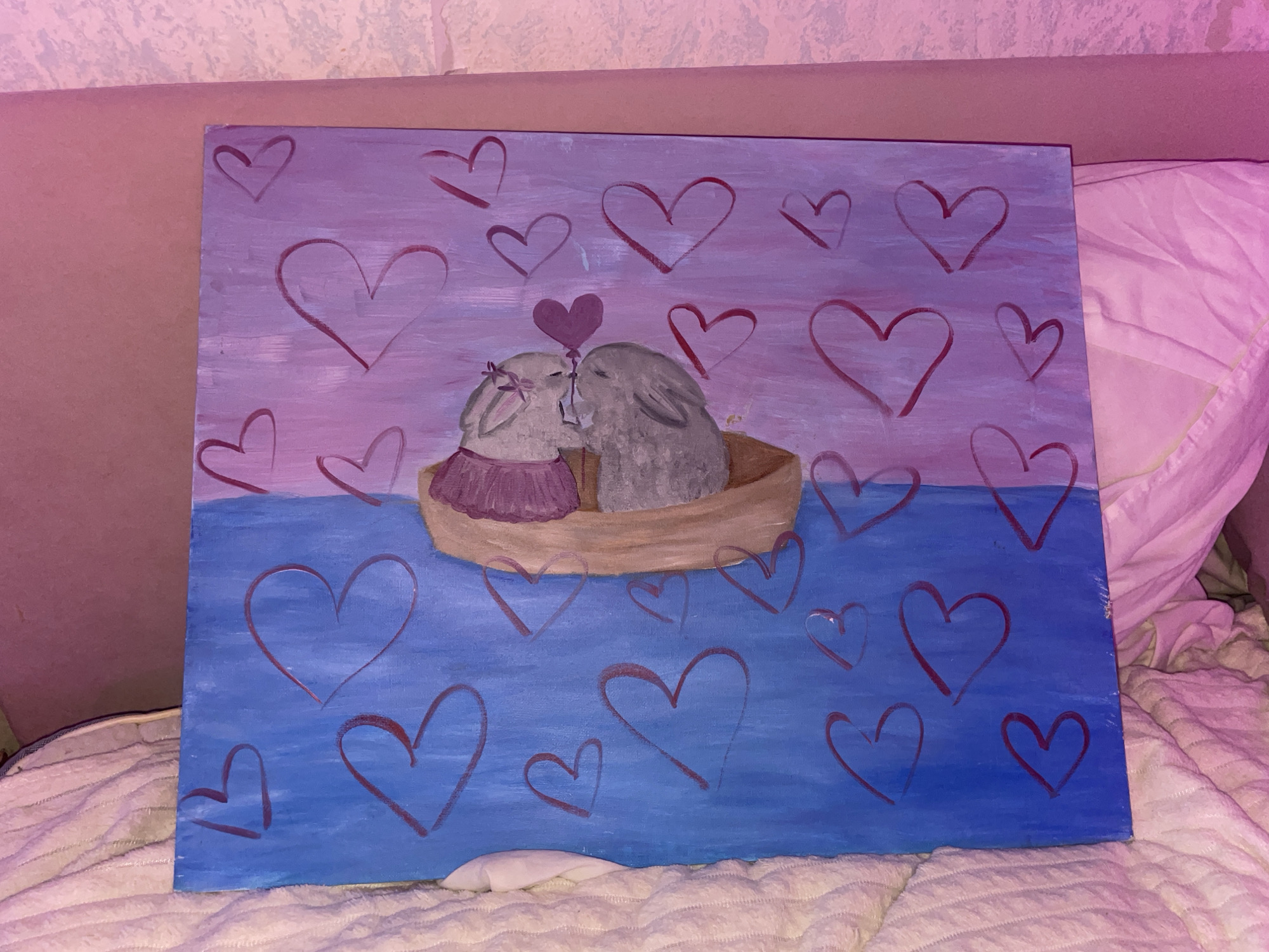 The media file displays a rectangular painting depicting a whimsical and romantic scene. The artwork features two cartoon-style bunnies in a small, light brown boat. The bunny on the left is lighter in color, adorned with a purple dress and what appears to be a floral headpiece, while the bunny on the right is grey. They are posed as if kissing or nuzzling, with a small dark purple heart-shaped balloon seemingly positioned between their heads.
The painting's background is divided into two horizontal sections: an upper part in shades of lavender and light purple, representing the sky, and a lower part in varying tones of blue, symbolizing water. Both the sky and the water are decorated with numerous hand-drawn outlines of red hearts in various sizes, enhancing the theme of love. The style appears to be acrylic or oil, with visible brushstrokes, suggesting a handmade quality.
The painting is photographed resting on a textured, off-white or light beige blanket or comforter. Behind it, a muted pinkish-brown surface, likely a wall or a piece of furniture, is visible. The entire scene in the photograph is bathed in a distinct pinkish-purple ambient light, which subtly alters the perception of the painting's original colors and creates a warm, intimate atmosphere. This lighting suggests an indoor setting, possibly during the evening, in Votkinsk, Russia. There is no visible text on the painting.