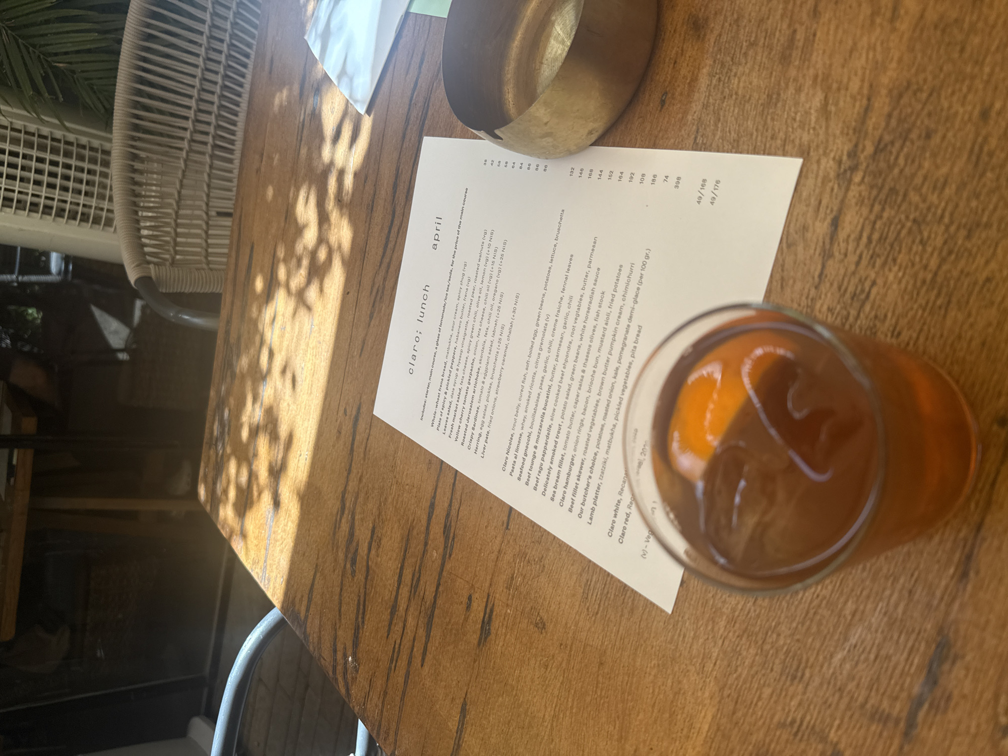 The image shows a wooden table at the Claro restaurant, as indicated by the printed "claro; lunch" menu. Claro is located in the Sarona complex in Tel Aviv, Israel. A menu titled "april" is visible with food options and prices. A glass filled with an amber-colored liquid, presumably a beverage, sits on the table. Next to the menu, there is a small, unidentifiable metallic container. The table is lit by sunlight casting leaf shadows. The table is partially surrounded by what appears to be a white wicker chair. A second chair is visible in the background.