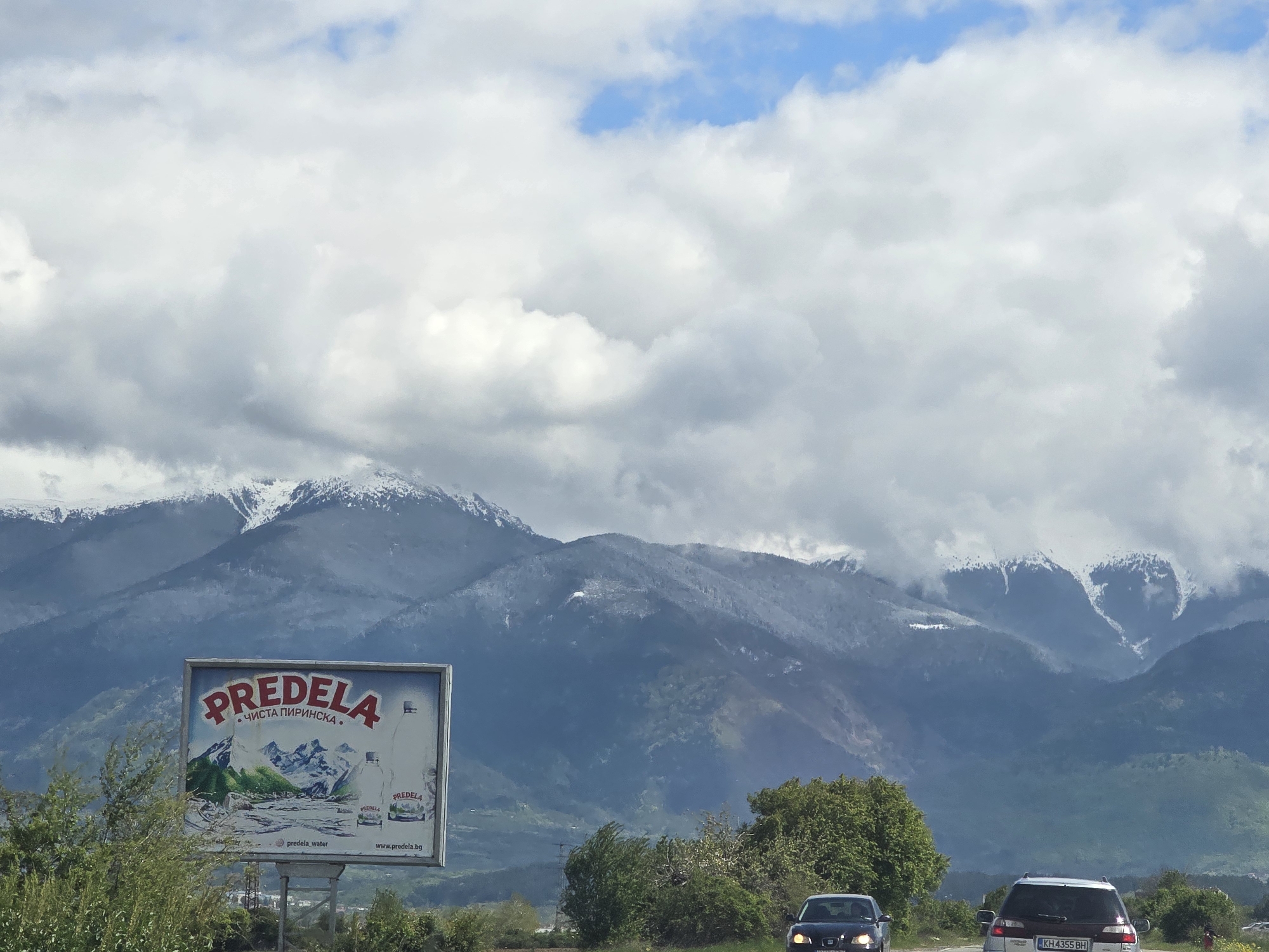 The image presents a striking mountainous landscape under a cloudy sky, likely in Piperevo, Bulgaria, as indicated by location cues. Snow-capped mountain peaks stretch across the background, partially obscured by large, white and grey clouds, with occasional patches of blue sky visible. This suggests a cool weather day, possibly in spring or autumn, given the presence of both snow and green vegetation.
In the foreground and mid-ground, a paved road winds through the scene. A prominent billboard stands on the left side of the road, advertising "PREDELA" brand water. The billboard features an illustration of mountains and two water bottles, alongside the Bulgarian text "ЧИСТА ПИРИНСКА" (Pure Pirin), which refers to the Pirin mountains in Bulgaria. Further text includes an Instagram handle "@predela_water" and a website "www.predela.bg".
Two cars are visible on the road. A dark sedan with its headlights illuminated is in the center foreground, while a silver SUV/wagon is seen further back on the right. The SUV/wagon bears a Bulgarian license plate, "KH 4355 BH," reinforcing the location. Lush green trees and bushes line the roadside, providing a contrast to the rugged mountains and the overcast sky. The scene depicts a journey through a beautiful, natural environment during daylight hours.