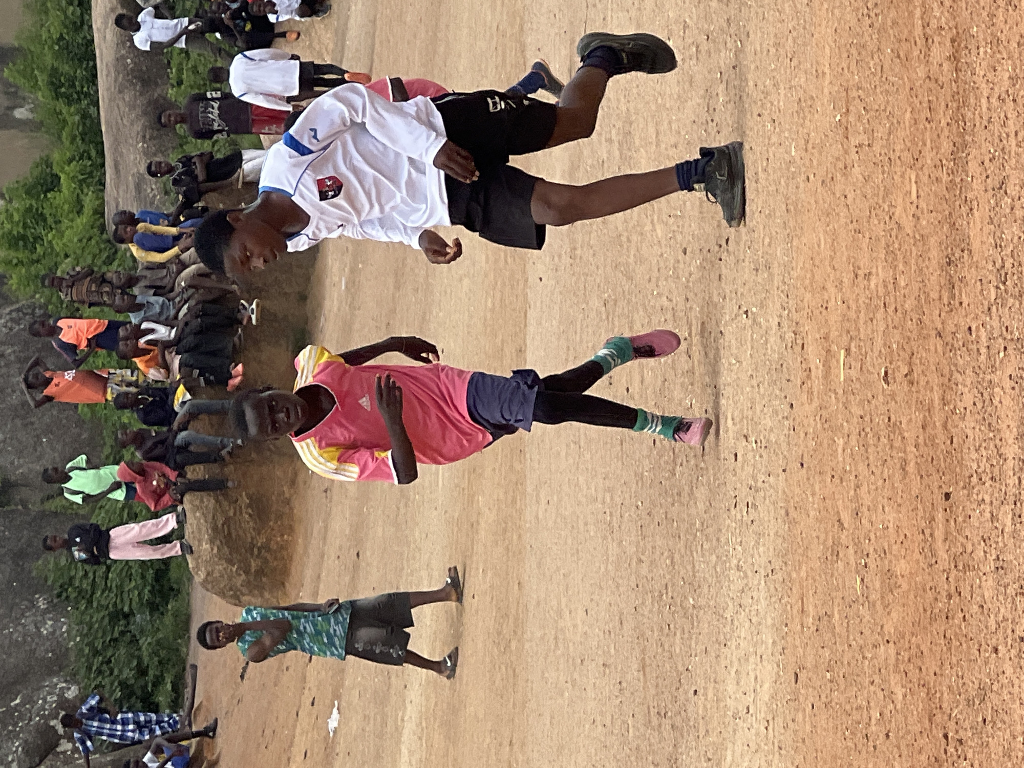 In Jos, Nigeria, a group of young boys are engaged in a soccer game on a dirt field. The foreground features two boys in motion: one in a pink shirt and dark shorts, the other in a white long-sleeved shirt and black shorts. Both are actively running, suggesting they are part of the game. In the background, a crowd of other boys and young men are observing from a rocky outcrop and sitting on the ground. The environment is characterized by dry, reddish-brown dirt and sparse vegetation, typical of an urban open space in Jos. The lighting indicates it is daytime, with overcast skies. The overall scene depicts a casual sporting activity amidst a community gathering.