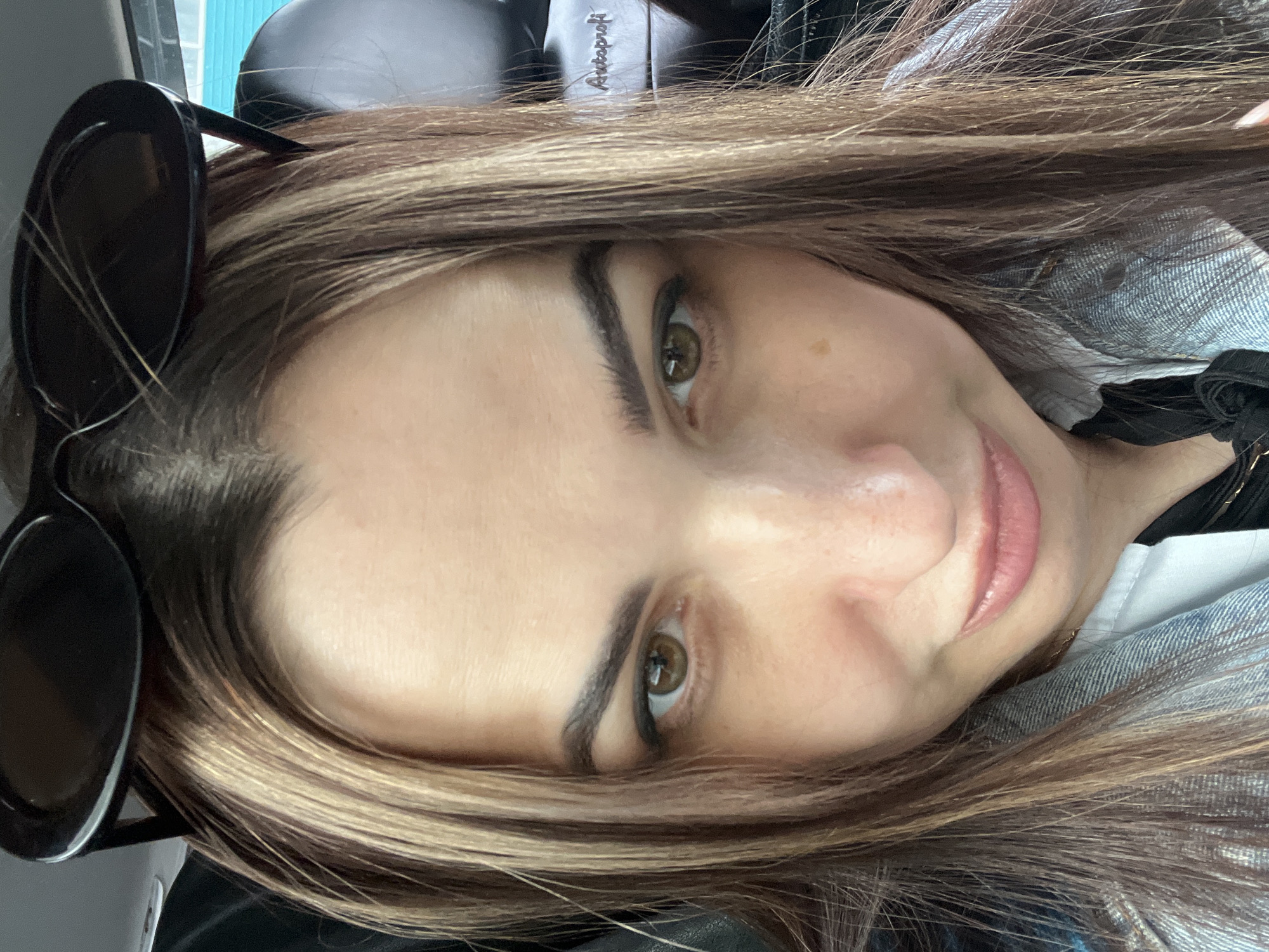 A close-up, angled selfie photograph shows a woman with medium-length, highlighted brown hair. She has dark eyebrows, light skin, and light brown eyes. She is smiling slightly. Sunglasses are perched on her head. She is wearing a denim jacket over a white shirt or undershirt. The background includes a headrest and what appears to be a car interior. The location is unknown.