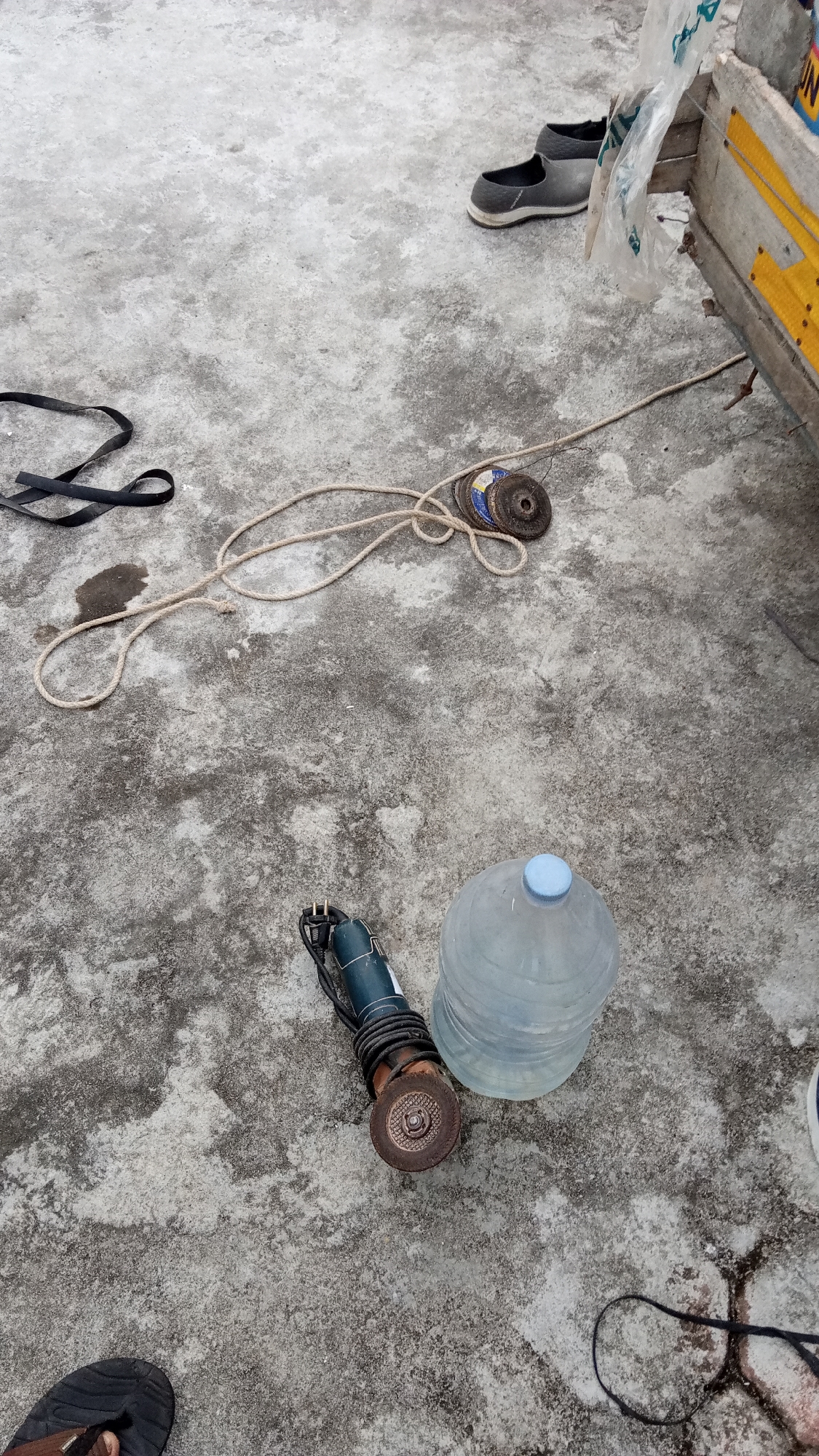A high-angle medium shot captures an outdoor scene on a rough, concrete surface. In the upper right quadrant, a pair of grey slip-on shoes are placed next to a wooden structure with yellow accents and a clear plastic bag. Below and to the left of the shoes, a coiled rope lies on the concrete, with a small stack of metal discs positioned near its center. To the left of the rope, a black, flat strap is also present. In the lower half of the image, a small, blue-handled grinder with a metal cutting disc is situated next to a large, clear plastic water jug with a blue cap. In the bottom left corner, a person's left foot wearing a black flip-flop is visible. The scene appears to be on a ground level surface in Bireuen, Indonesia, potentially a workshop or outdoor storage area.