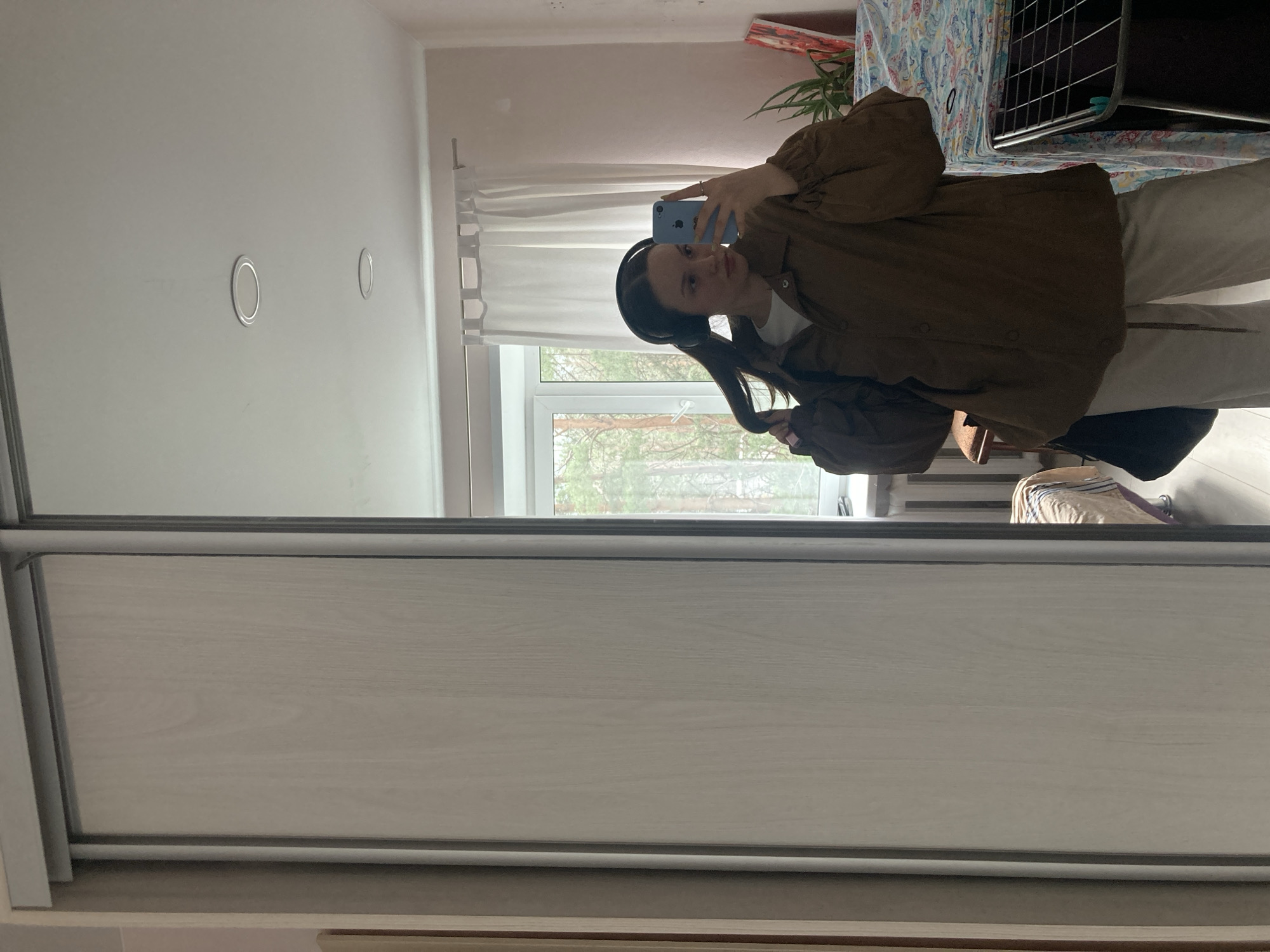 A woman with dark hair and wearing headphones is taking a selfie in a mirror. She is wearing a light-colored shirt and pants, and a brown jacket. In the background, there is a window showing trees, a curtain, a plant, and a drying rack. The mirror is part of a wardrobe with light-colored doors. Based on the available information and visual cues, there is not sufficient data to specify the location in the city of Chaikovsky, Russia.