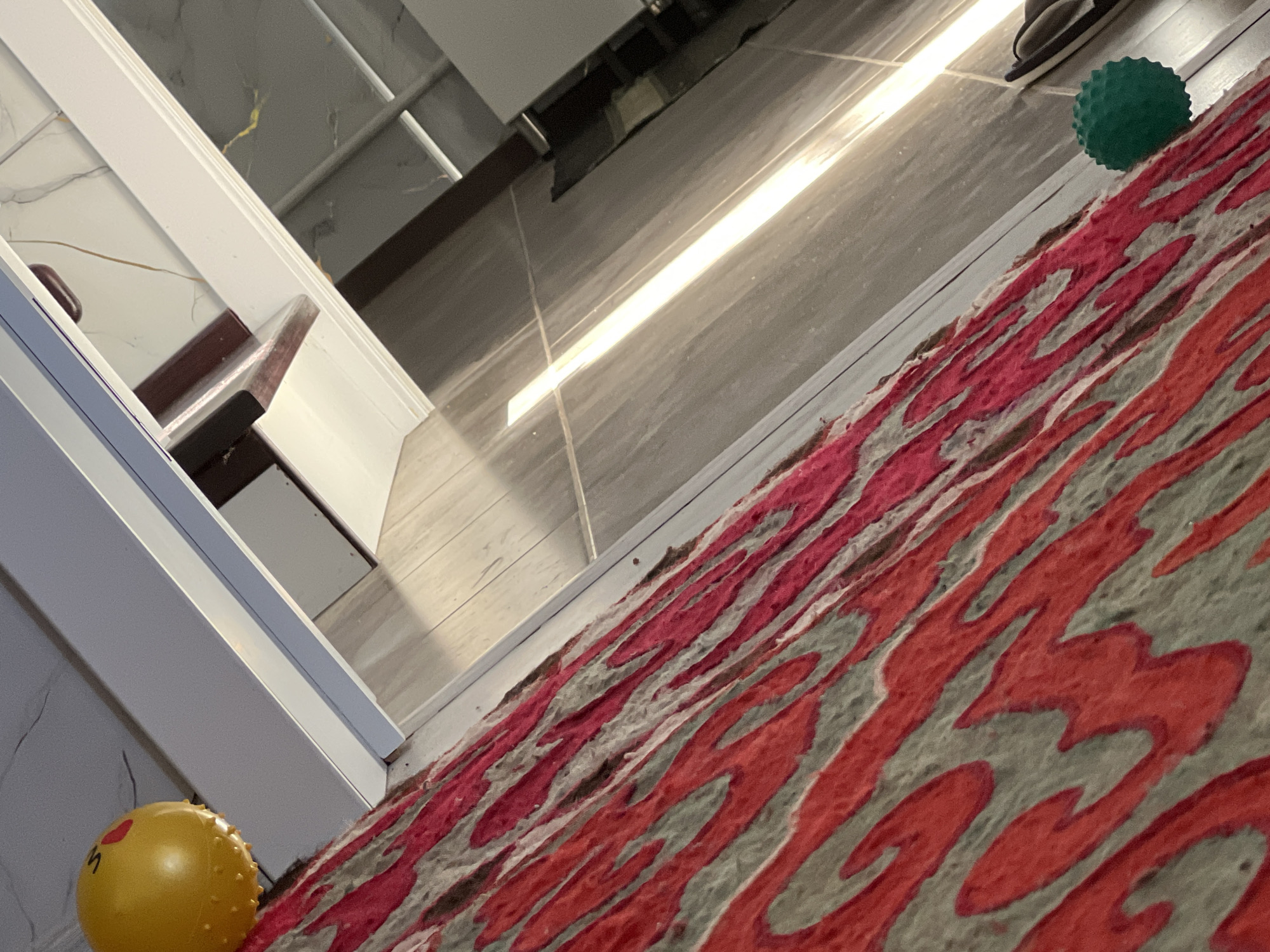 The image depicts an indoor scene, possibly a hallway or a room with a tiled floor and a white trim along the wall. A rug with a red and gray abstract pattern lies diagonally across the foreground. Two toys are visible: a yellow ball with a red emoji-like face on the left, and a small green textured ball on the right. The lighting suggests a bright light source, possibly from a window or an overhead fixture, casting a long, illuminated stripe across the floor. The overall composition is at an angle, giving a skewed perspective of the environment. There is no indication of the location being in Бирдик, Kyrgyzstan.