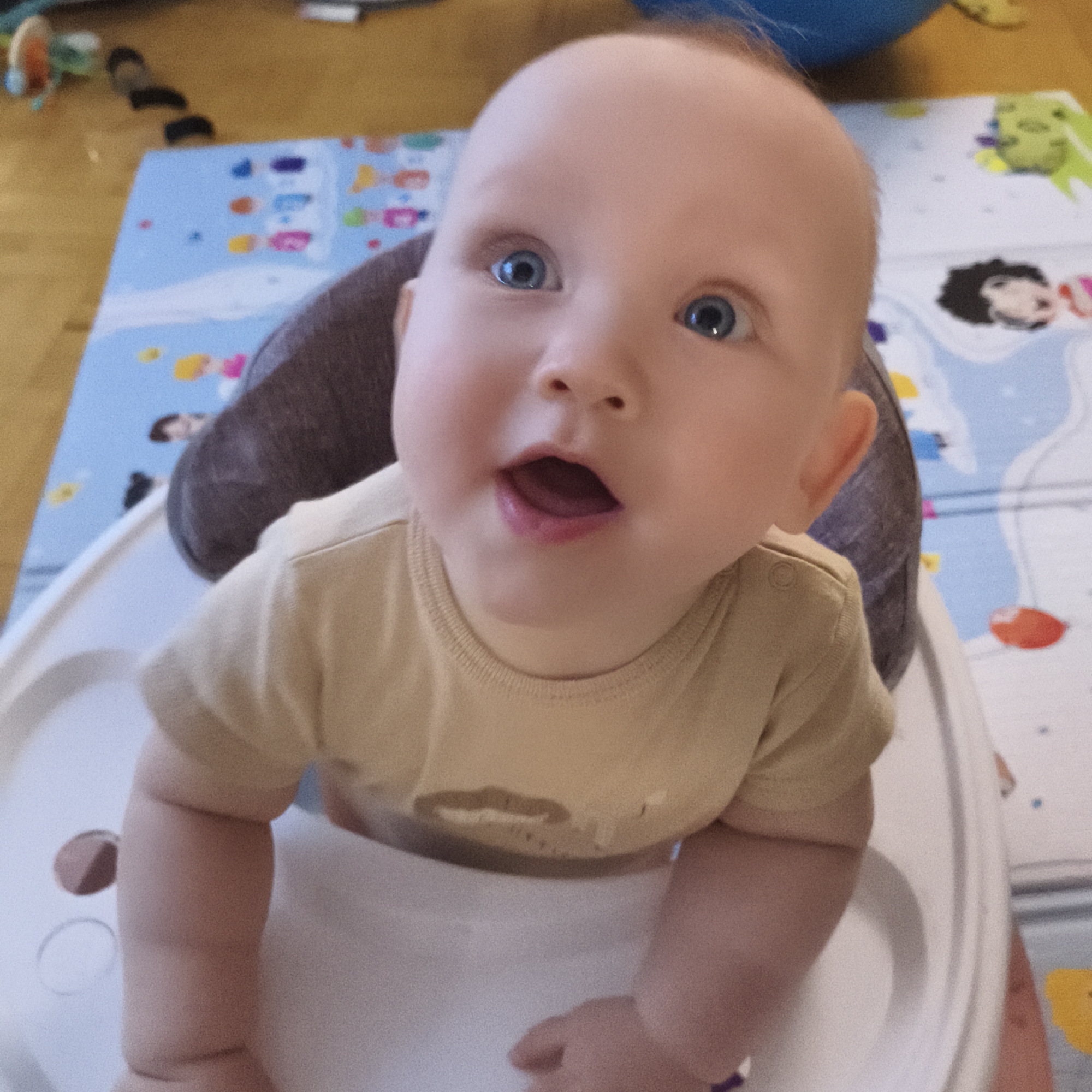 An eye-level, close-up photograph captures an infant seated in a light-colored baby seat with a dark padded insert, in an indoor domestic setting in Chaikovsky, Russia. The baby, appearing to be several months old, has fair skin, light-colored hair, and striking bright blue eyes that are wide open and focused directly towards the camera. Its mouth is slightly open, revealing a pink tongue, suggesting a moment of surprise, curiosity, or vocalization.
The baby is wearing a plain light yellow or beige short-sleeved t-shirt. Visible around the baby seat and extending into the background is a colorful, light blue playmat adorned with various blurry cartoon-like figures, shapes, and what appear to be parts of letters or numbers, including a depiction of a head with dark curly hair. Beneath and beside the playmat, a warm-toned wooden floor is visible. In the upper left background, indistinct colorful toys are scattered on the wooden floor, and a blurry blue object, possibly a ball, is partially seen in the upper right.
The lighting suggests it is daytime. The infant's expression is one of alertness and engagement, conveying a sense of innocent wonder. The photograph has a shallow depth of field, with the baby's face sharply in focus while the background elements are softer. There is no clearly legible text visible in the image.