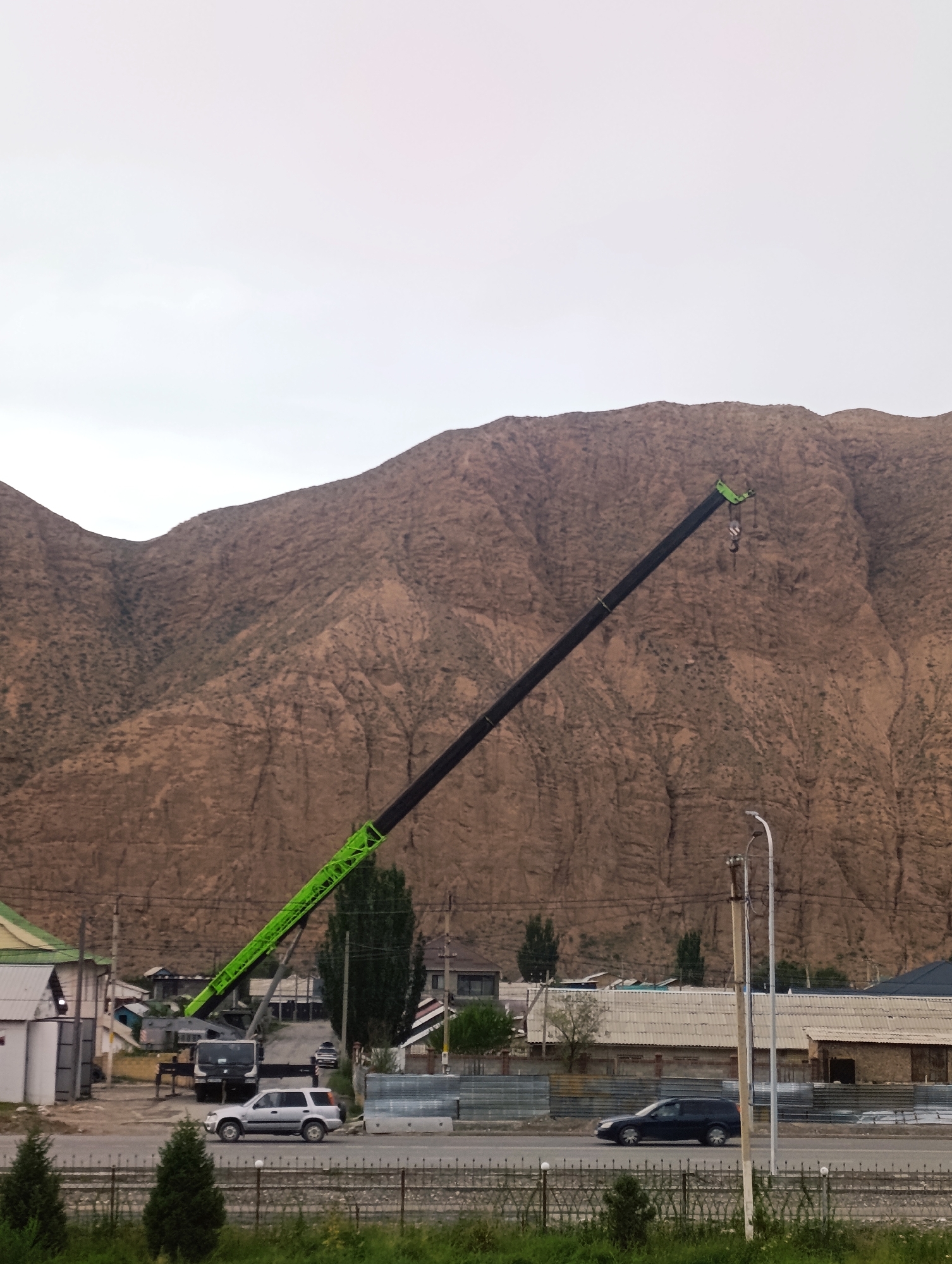 An outdoor, eye-level, full shot captures a large crane with a long black and green boom angled across a brown, rocky mountain in Naryn City, Kyrgyzstan. The crane is positioned in front of a cluster of low-rise buildings and a road. A silver SUV is parked on the side of the road in the foreground. A dark-colored sedan drives on the road. A street light stands next to the road. The sky is overcast and pale.