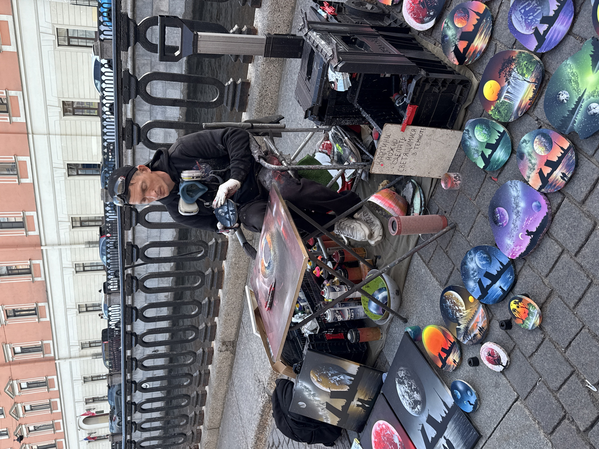 A street artist is creating spray-painted art on a cobblestone sidewalk next to a canal in Saint Petersburg, Russia. The artist, wearing a black hoodie, goggles on his forehead, and a respirator mask around his neck, sits in a folding chair. He is working on a small, portable easel with a circular painting. Surrounding the artist are various spray cans, brushes, and finished artworks displayed on the ground. These artworks are predominantly oval-shaped and feature celestial or natural landscapes with silhouettes. Behind the artist, a black metal fence separates the sidewalk from the canal, and in the background, a building with a salmon-pink facade and numerous windows is visible. A sign on an easel reads "КАРТИНЫ МОЖНО СДЕЛАТЬ СВЕТЯЩИМИСЯ В ТЕМНОТЕ" (Paintings can be made glowing in the dark).
