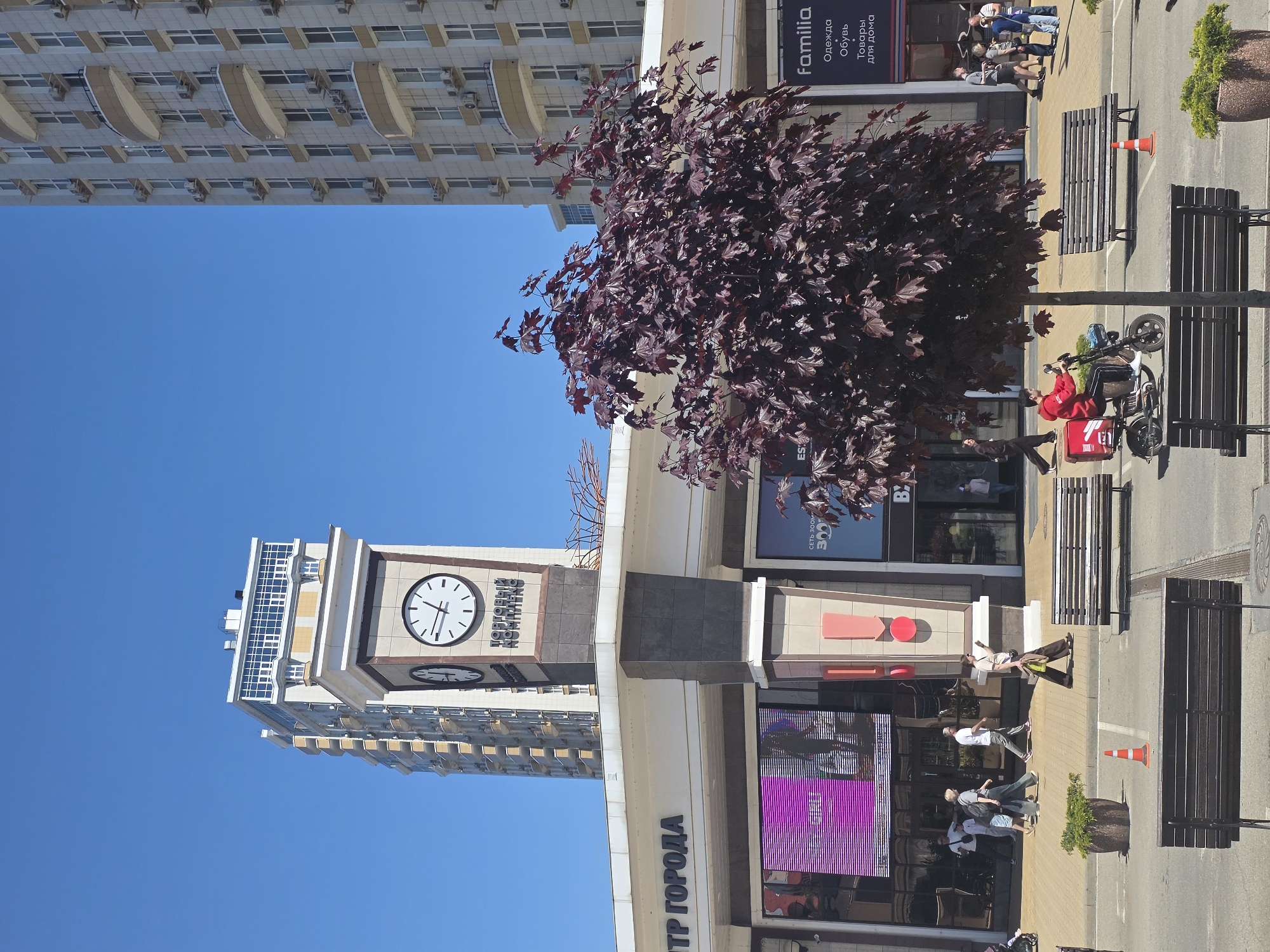 The image shows a street scene in Krasnodar, Russia. Dominating the left side is a tall building topped with a clock tower labeled "ТОРГОВЫЙ КОМПЛЕКС". The sky is clear blue. In the foreground, there's a shopping complex with shops and illuminated displays. A dark-leafed tree stands in front of the complex. People are walking along the paved sidewalk. There are benches along the sidewalk and an orange traffic cone on the street. A delivery person on a scooter with a red delivery bag is parked next to one of the benches. The overall setting is a busy urban area.
