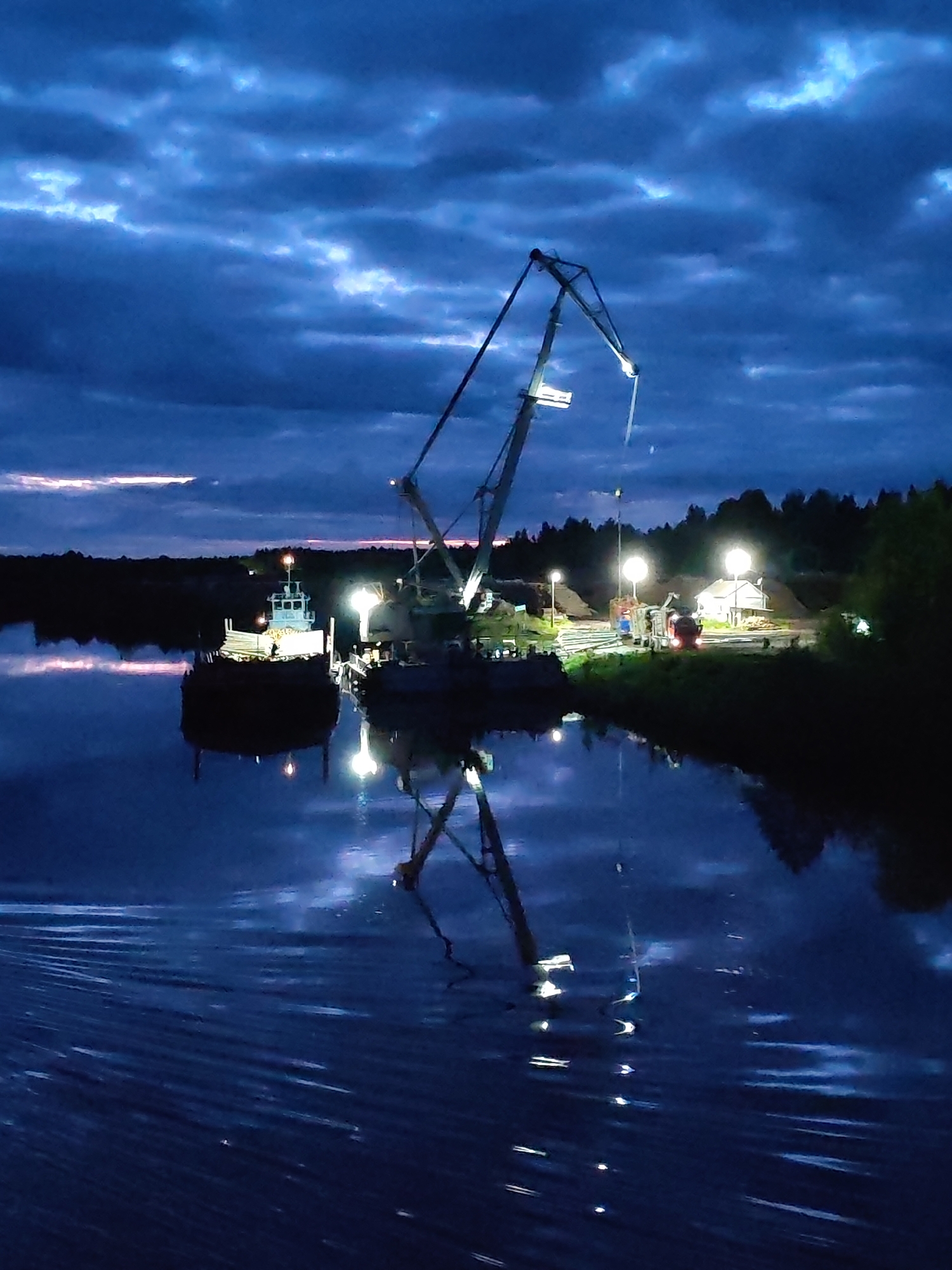 A nighttime view of a river or large body of water in Annensky Most, Russia. A large crane is visible on the bank of the river along with a tug boat or small vessel. Lights are shining on the land nearby, possibly indicating a construction or industrial site. The sky is cloudy. The water reflects the light from the crane and the other sources of illumination. Ripples on the water suggest a slight breeze.