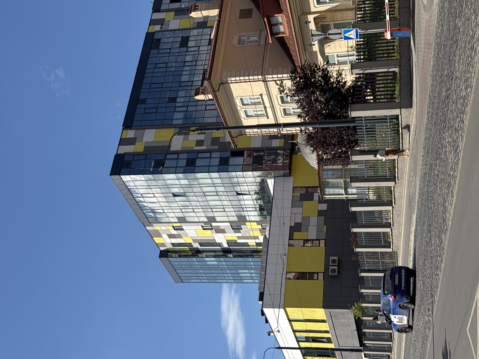 The image depicts a street scene in Lviv, Ukraine, under a clear blue sky. The primary subjects are modern buildings with distinctive glass facades featuring a geometric pattern of blue, yellow, and gray panels. Adjacent to these is a more traditional, lighter-colored building with a red tiled roof and visible windows.
In the foreground, a blue car with "Bolt" branding is parked on a cobblestone street. Two people are visible near the car, with one appearing to be either entering or exiting the vehicle. Further along the street, a person and a dog are walking. There are also traffic signs and a pedestrian crossing visible.
The scene appears to be set during the daytime, with strong sunlight casting shadows. The weather is clear and sunny. The presence of the Bolt car and the architectural styles suggest an urban environment. The image is taken from a slightly elevated perspective looking down at the street and buildings.