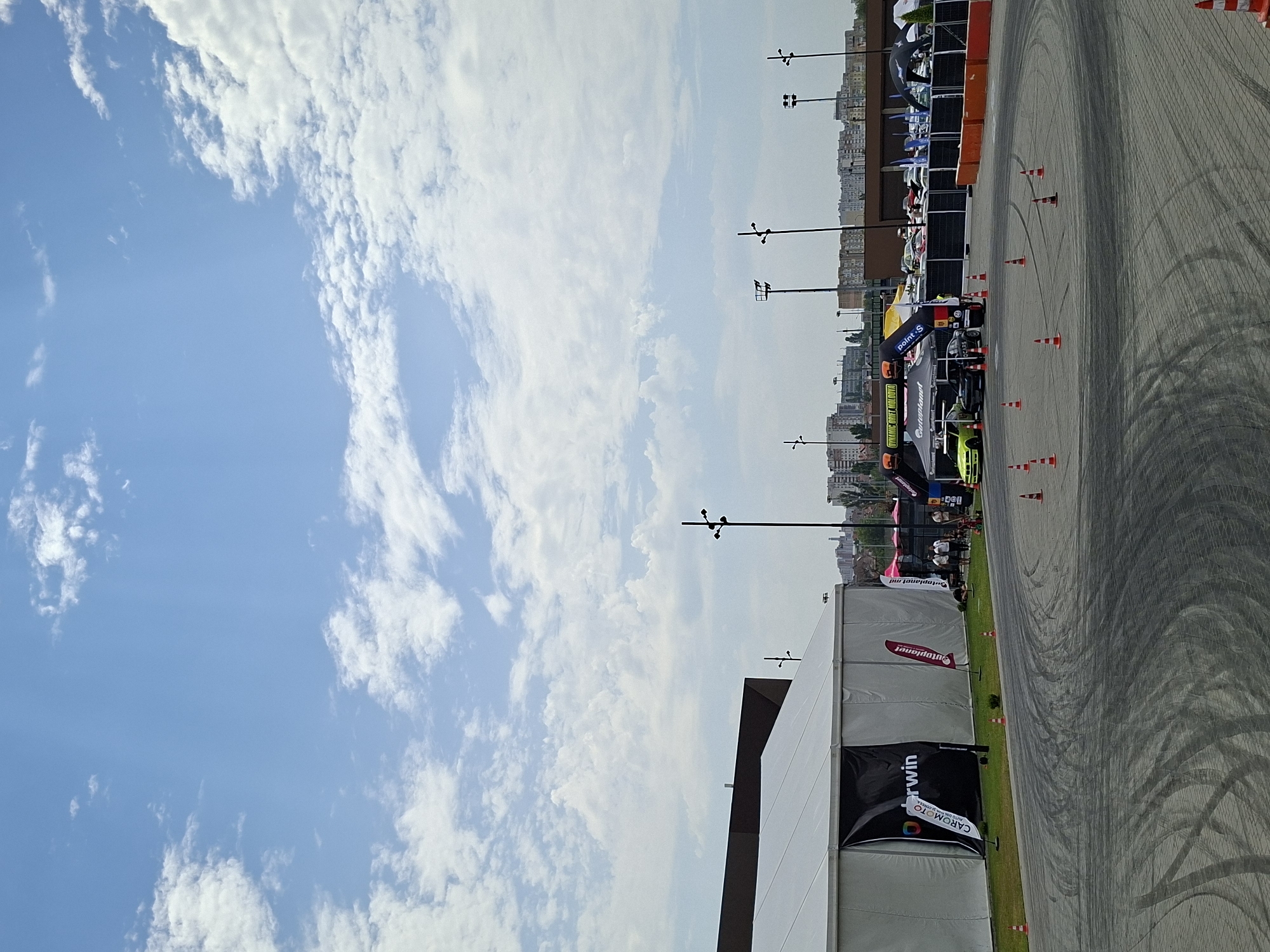 This is a wide, eye-level shot of an outdoor asphalt track with tire marks from drifting, likely at the Stăuceni Speedway in Stăuceni, Moldova. In the background, a large white tent structure stands on the left, displaying a banner with the text "Darwin." To the right of the tent, a yellow and black archway reads "DYNAMIC DRIFT MOLDOVA." Several tents and booths are set up behind the archway, with cars visible in the distance. Orange and white traffic cones are placed around the track to mark boundaries. The sky above is blue with scattered white clouds. The scene appears to be set during daylight hours.