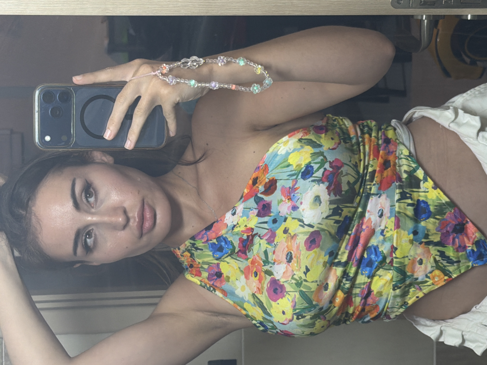 A woman is taking a selfie in a bathroom, holding a smartphone in her right hand. She is wearing a floral-print, one-shoulder top and white shorts. The smartphone has a clear case and a beaded phone charm with flowers and beads. Her left arm is raised, and her hair is partially pulled back. The background shows a mirror, part of a door frame, and a yellow object that appears to be a child's toy. The scene appears to be indoors in a domestic setting, consistent with a residence in Tersefanou, Cyprus.