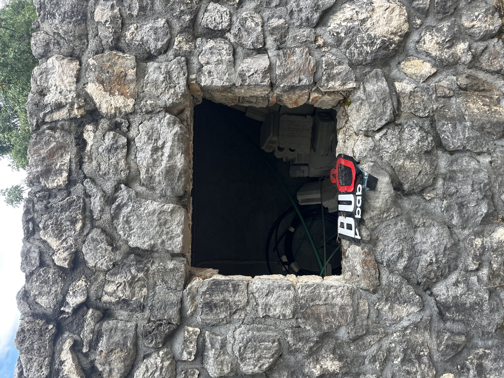 The image shows a rough-hewn stone structure with a rectangular opening. A black and red sign with white lettering that reads "BU bab" is affixed to the stone just below the opening. Inside the dark opening, green hoses and what appear to be white construction materials or architectural elements are visible. Trees with green foliage are present at the top of the frame, suggesting an outdoor location. The structure's rough, unrefined construction implies it may be part of an older or more rustic building, possibly in a less developed area. The stone wall is made of irregularly shaped gray stones cemented together. The sky is visible at the very top of the image and appears cloudy. The scene does not contain any people or discernible action.