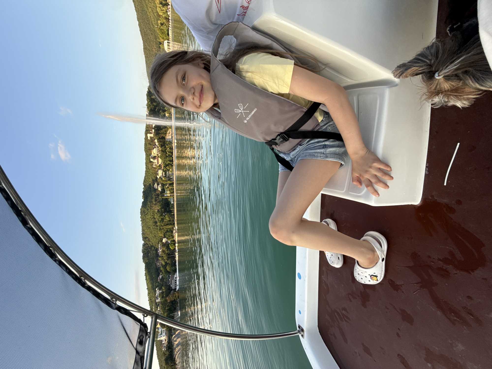 A young girl with light brown hair, wearing a yellow t-shirt and denim shorts, sits on a boat with a life vest on. She is smiling at the camera. To her right, a small dog with long, dark fur is partially visible. The boat is on a body of water, with a green, tree-covered hill and buildings in the background. A large fountain sprays water into the air in the distance. The sky is clear and blue, suggesting it is daytime. The overall mood is happy and relaxed. A logo on the girl's life vest appears to read "BUOYABRAU."