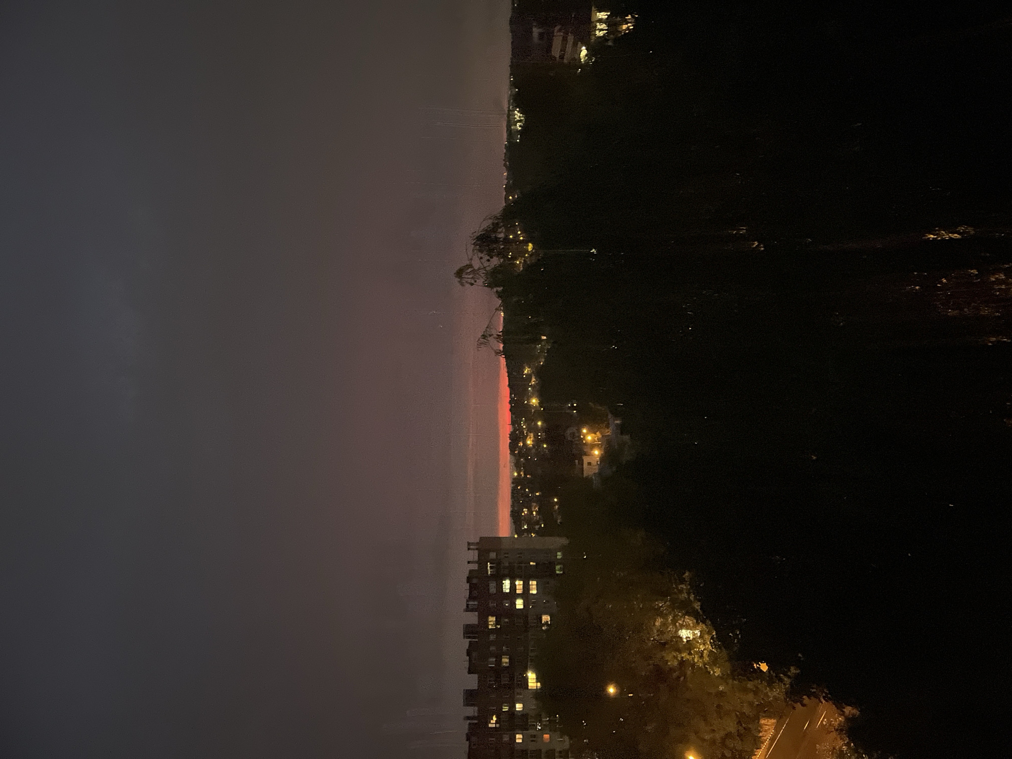 A dusk cityscape is depicted with a deep purple and pink gradient sky. A large apartment building with illuminated windows is visible on the left side of the frame, with trees in the foreground obscuring the lower half of the image. In the distance, a sprawling network of smaller buildings and streetlights creates a warm glow. The overall atmosphere is serene and tranquil. The scene is captured from a high vantage point, likely a window or balcony, looking out over the urban landscape.