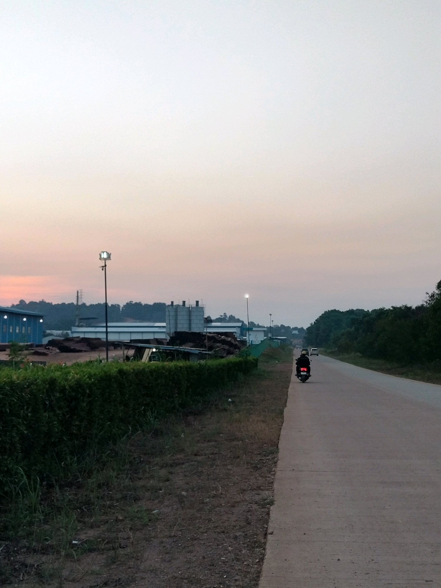 The image depicts an outdoor scene on a paved road with a motorcycle and two cars. The sky is overcast with a soft, pastel gradient of colors from light blue at the top to a peachy hue at the horizon. On the left side of the road, there is a dense green hedge running parallel to the road. Beyond the hedge, industrial buildings with blue and white facades are visible, along with stacks of what appears to be lumber or raw materials. A tall lamp post with a bright light is positioned near the hedge. In the distance, more industrial structures and trees can be seen. On the right side of the road, a dense line of trees forms the background. A person is riding a black motorcycle on the road, with its red taillight illuminated. Two cars are visible further down the road in the same direction as the motorcycle. The scene appears to be set in an industrial area on the outskirts of Batam, Indonesia, likely at dusk or dawn due to the lighting conditions and the soft sky colors.