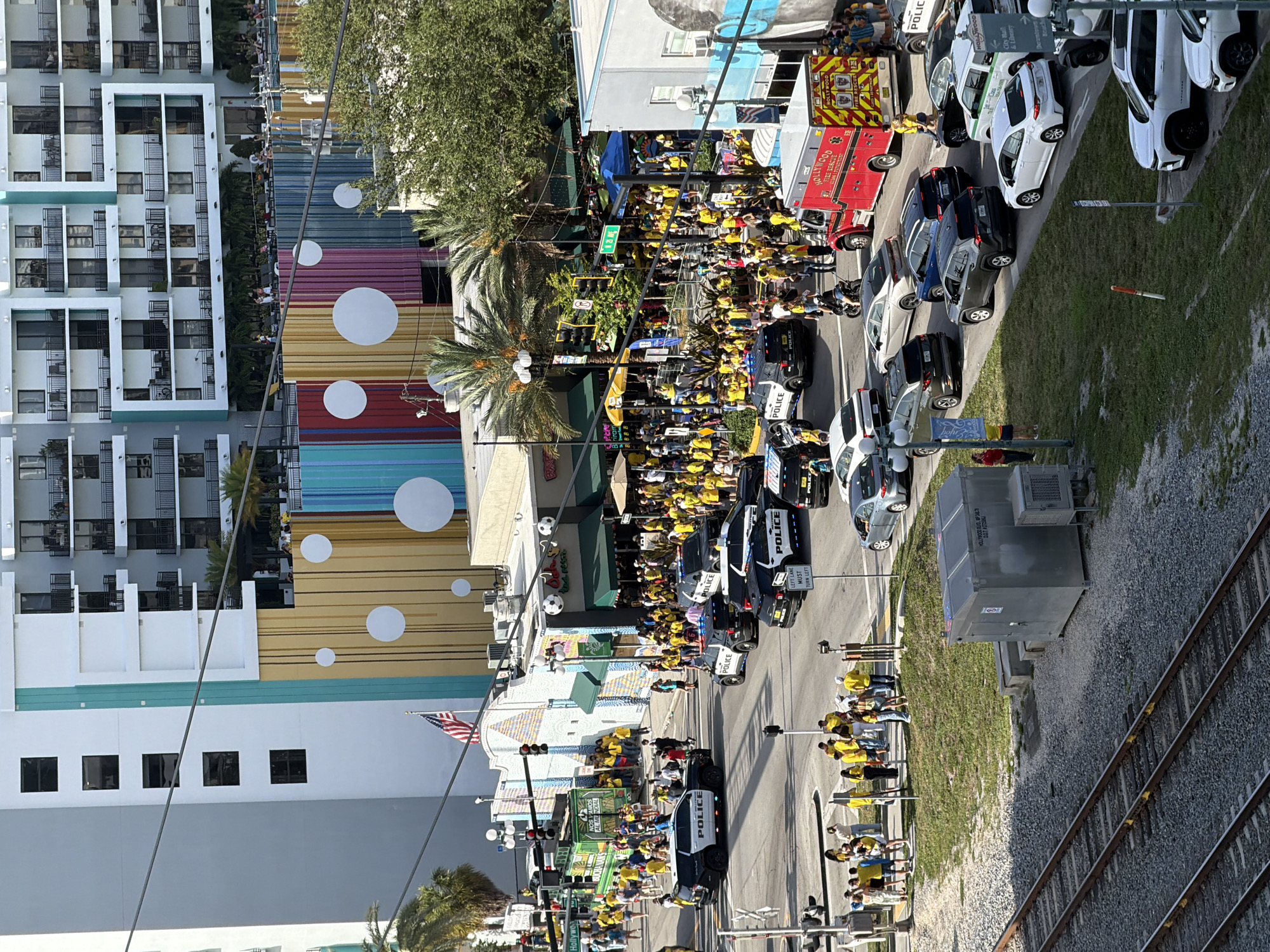 This is an elevated, eye-level, full shot of a street scene in Hollywood, Florida, USA. A large crowd of people, many wearing yellow, are gathered on the sidewalk and street. Several police cars are present, some with their lights on, and an ambulance is visible. Buildings line the street, including a multi-story apartment building with balconies and a building with a colorful, striped facade. Traffic signals and streetlights are visible, as are palm trees and other greenery. In the foreground, train tracks run parallel to the street. The overall impression is of a bustling event with a strong police presence.