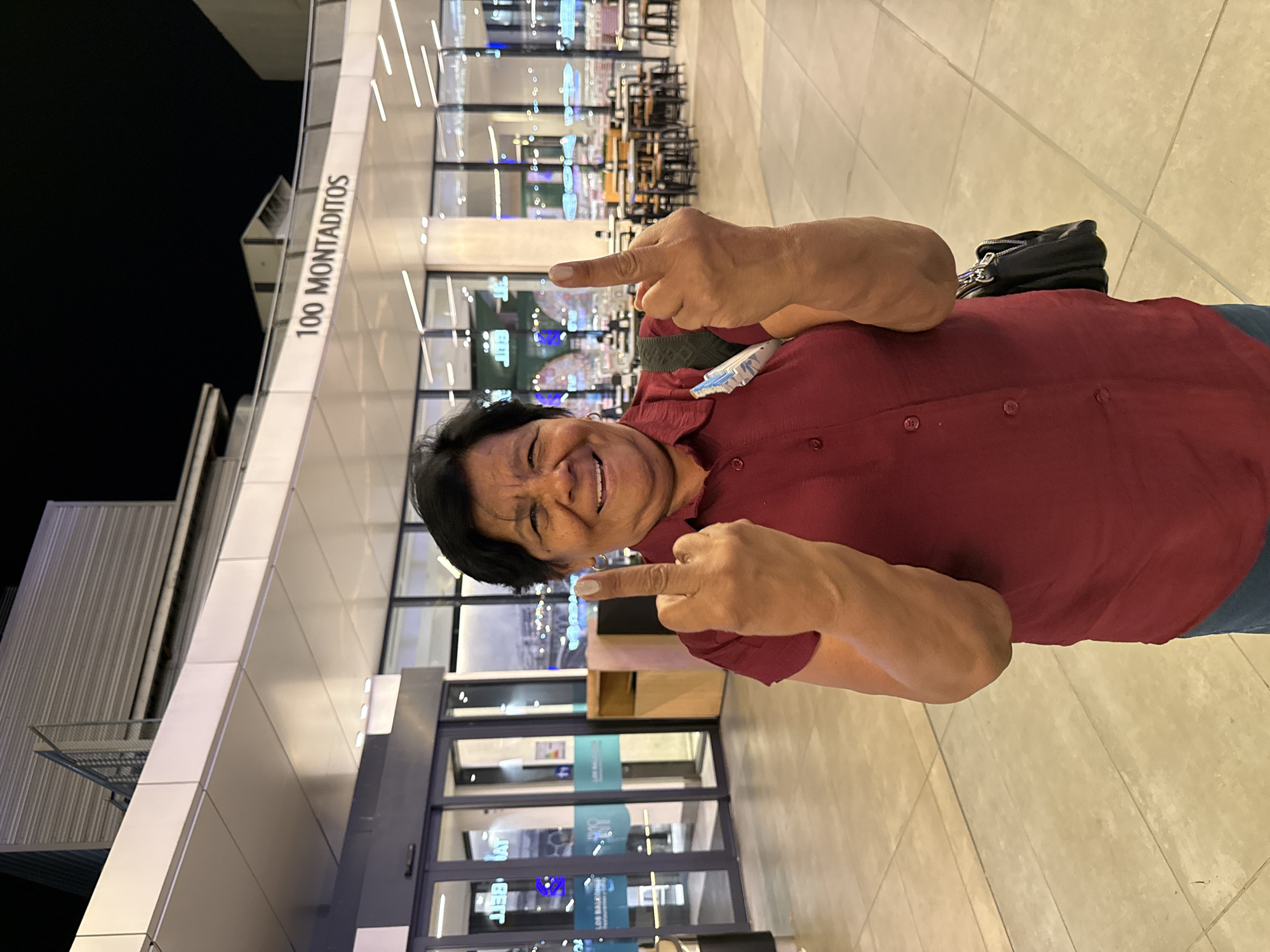 This media file is a vertical, full-body photograph of a person standing outside a modern building at night. The person, who appears to be of middle-aged or older, is smiling broadly and looking at the camera. They are wearing a maroon collared shirt and have their arms extended, with their index fingers pointing outwards, seemingly in a playful or defiant gesture. A black handbag is slung over their left shoulder.
Behind the person, the building features a sleek, metallic facade with geometric panels and large glass windows. The windows reveal interior lighting, suggesting the building is a commercial establishment, possibly a restaurant or retail space, with tables and chairs visible inside. The ground in front of the building is paved with light-colored tiles.
The text "100 MONTADITOS" is clearly visible in large, dark letters on a white panel on the building's exterior, indicating the name of the establishment. There are also some blue signs visible through the glass doors, though the text is largely illegible. The lighting suggests it is nighttime, and the scene appears to be well-lit. The overall mood is cheerful, with the subject's expression and gesture conveying a sense of fun and personality. The location context is Alcorcón, Spain.