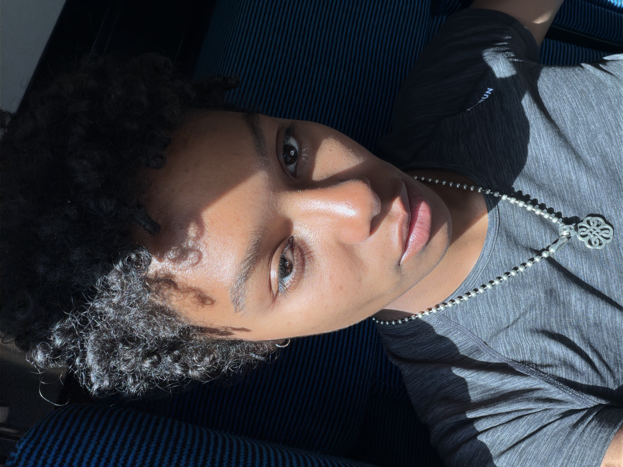 A person lies on a striped blue surface wearing a heather gray t-shirt and a silver beaded necklace with a pendant. The person has dark, curly hair. The person's skin has a sheen and is in the sunlight. The location is not specified in the image, therefore the city of Sainte-Eulalie, France cannot be confirmed.