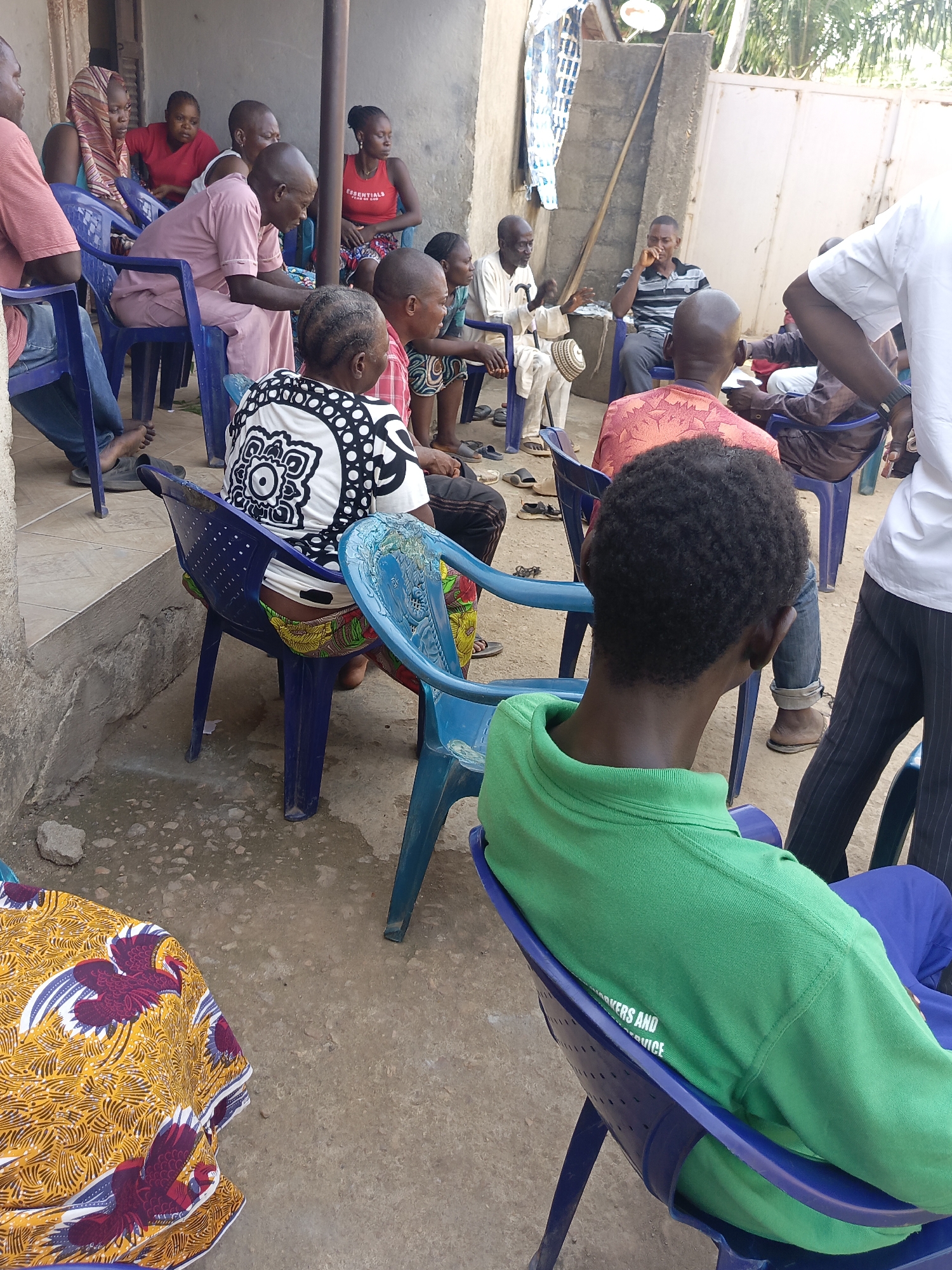 The scene shows a group of people gathered outside, likely in Akwanga, Nigeria. The group consists of men and women of varying ages, sitting on blue plastic chairs. Some individuals are dressed in traditional African clothing, while others wear more casual attire. The environment appears to be a courtyard or outdoor meeting space, with a concrete ground. There is a building or wall in the background, and some objects like a cloth with a floral print are on the ground. The people are engaged in what seems to be a meeting or discussion, with some individuals appearing to be speaking or listening attentively.