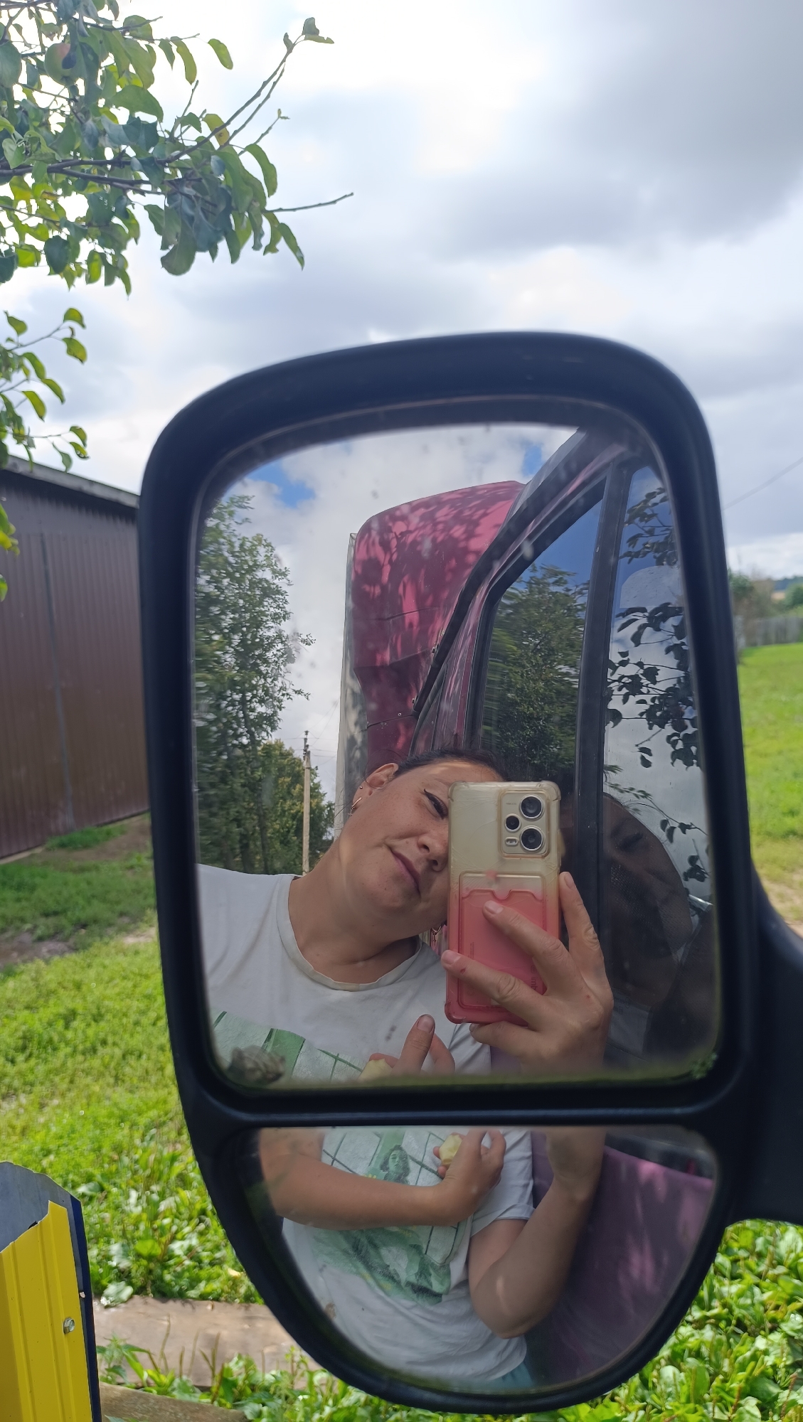 A person is taking a selfie in a truck's side mirror. The person is holding a pink-cased smartphone in their right hand, with their left arm bent and hand near their chest. They are wearing a white t-shirt. The mirror reflects a cloudy sky, green trees, and a brown building in the background. The person's face is partially visible, with their head tilted and eyes looking towards the phone. The truck's body, a dark red color with a distressed pattern, is also reflected in the mirror. The scene appears to be outdoors, possibly in a rural or semi-rural setting. The image does not provide any information about the location being Karsha, Russia.