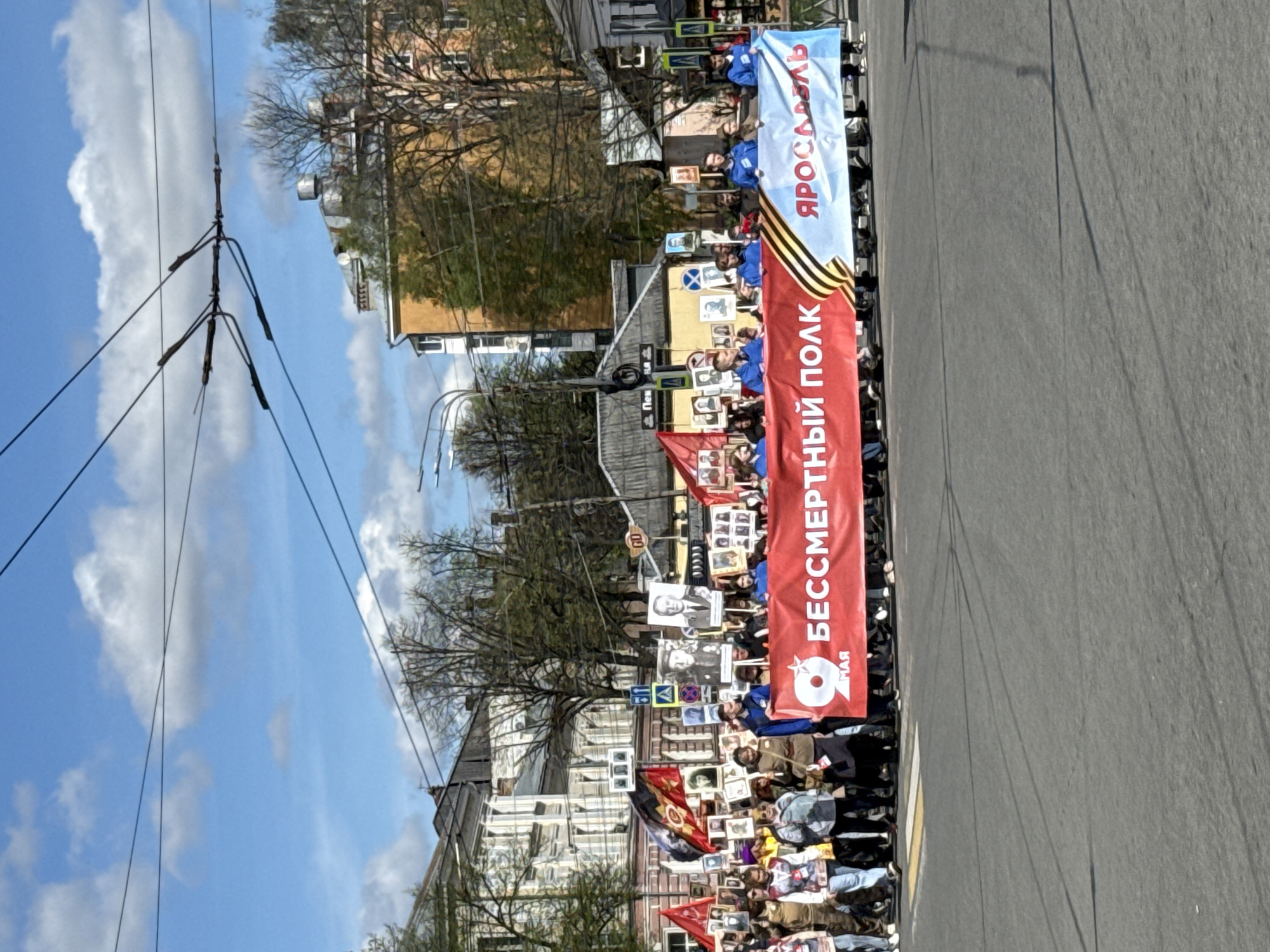 A daytime street parade proceeds through an urban environment. Numerous people march on the road, carrying banners, portraits, and flags. The banners contain Cyrillic text, including the phrases "Бессмертный Полк" (Immortal Regiment) and "Ярославль" (Yaroslavl). A tall red banner with a white inscription is prominently displayed in the middle of the parade. The marchers are diverse in attire. Background structures include buildings, trees, and utility poles. The sky is blue with scattered clouds.