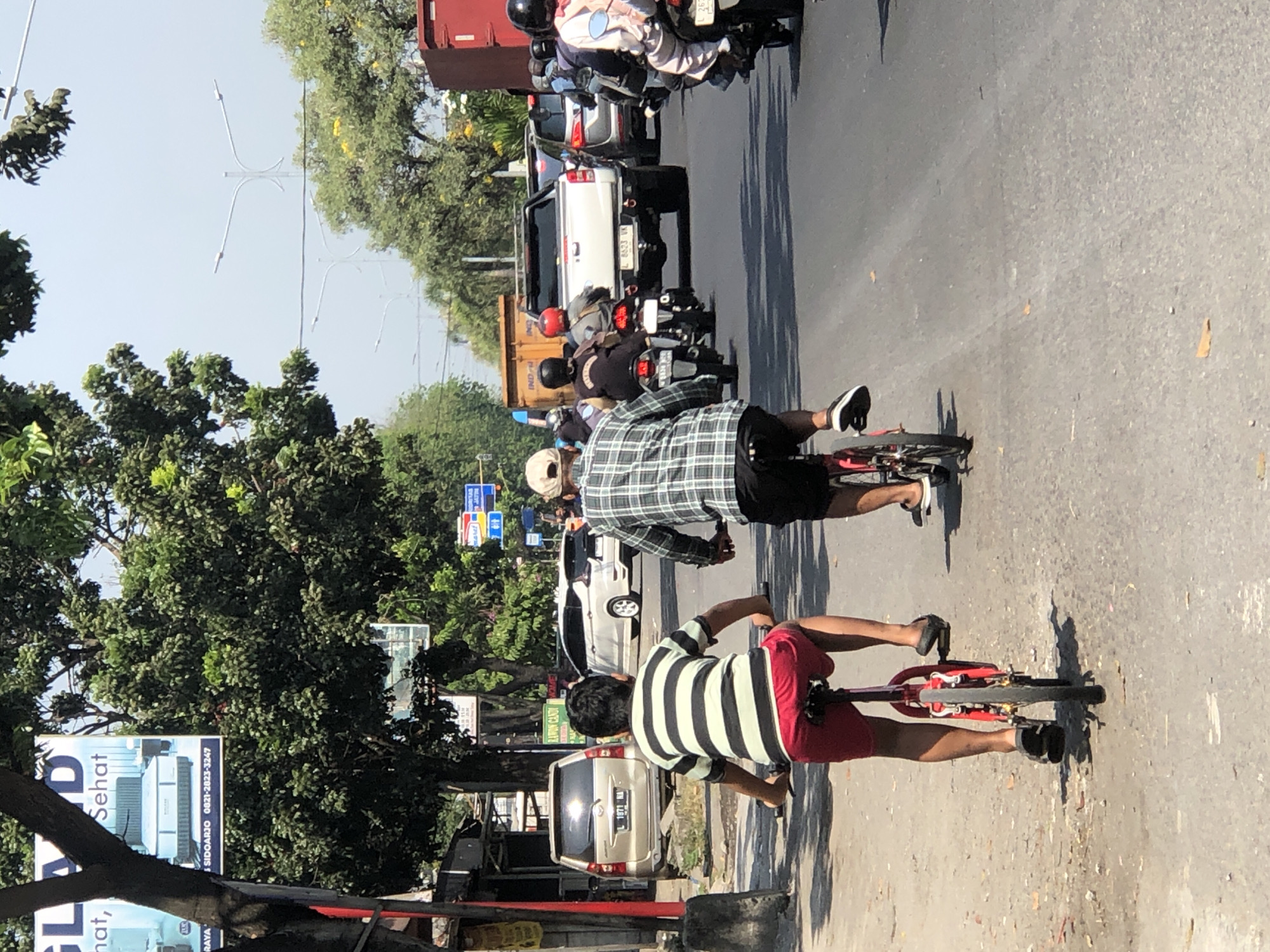 In Larangan, Indonesia, a sunny day sees traffic on a paved road. Two individuals on bicycles ride in the same direction on the right side of the road. The first cyclist, seen from behind, wears a black and white striped t-shirt and red shorts. They are on a red bicycle. Behind them, a second cyclist, also seen from behind, wears a plaid long-sleeved shirt and dark shorts, riding a bicycle. Motorcyclists and cars fill the lanes to their right, with a white pickup truck in the distance. The road is lined with trees and various advertisements, including one for mattresses. The scene captures a typical street view with a mix of vehicles and pedestrians.
