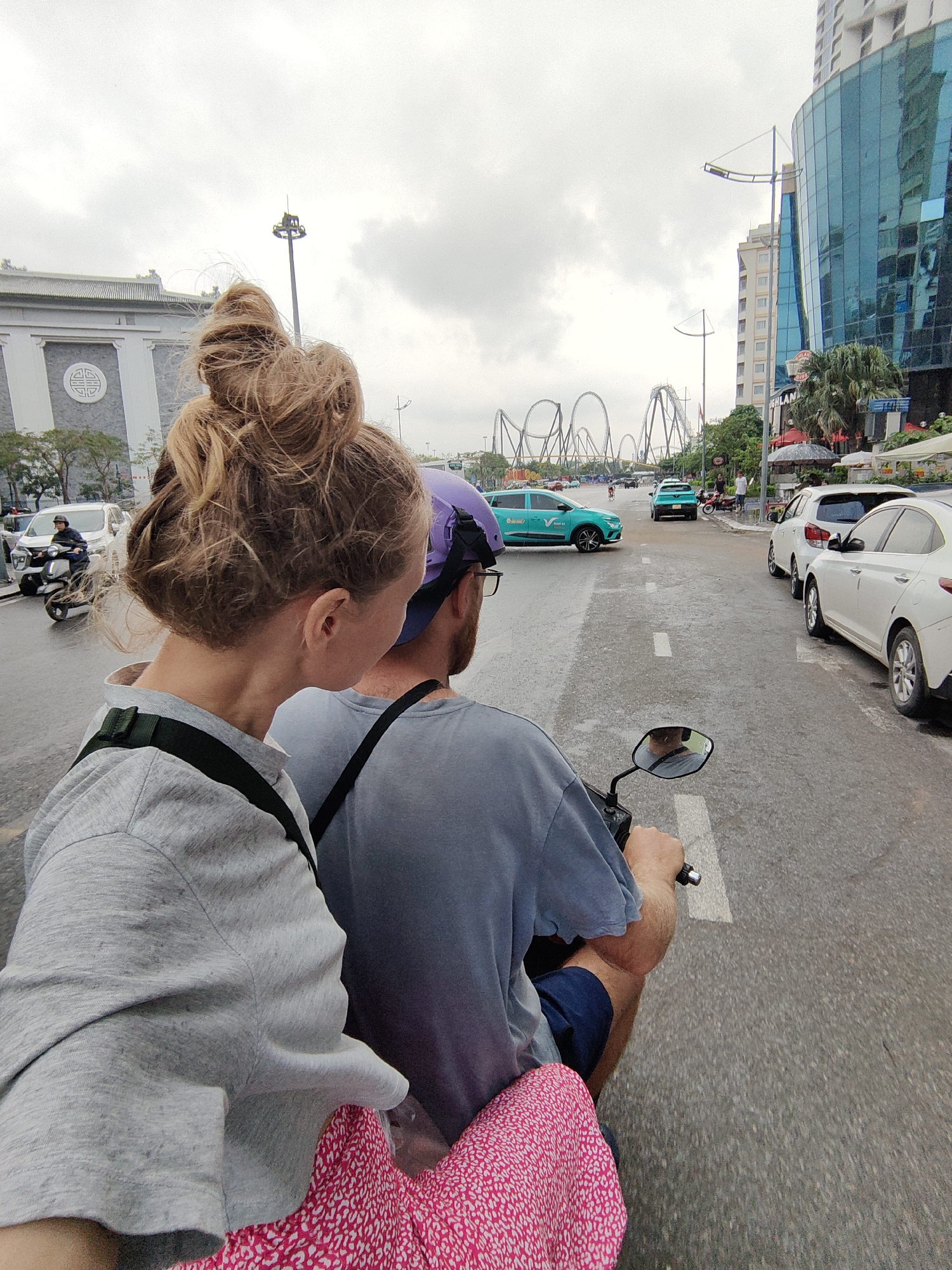 A couple is riding a motorbike down a wet road in Bai Chay Ward, Vietnam. The woman in the back has blonde hair styled in a bun and wears a gray sweatshirt. The man driving wears a purple helmet and a blue t-shirt. A pink bag is visible between them. The road is lined with parked white cars on the right, and other motorbikes and cars are visible ahead. A series of large, gray roller coaster loops are in the distance. Buildings are present on both sides of the street, including a modern blue glass building on the right. The sky is overcast.
