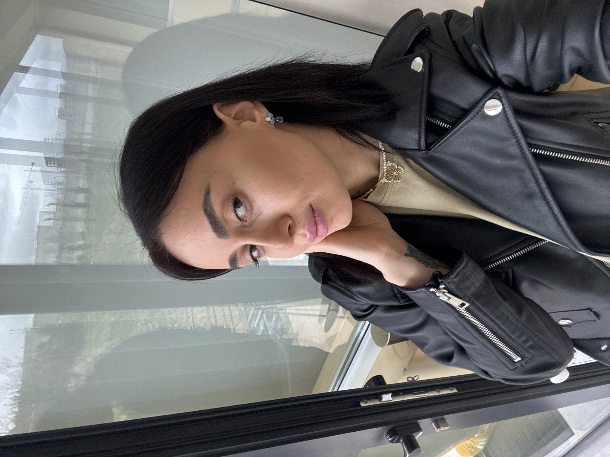 A female individual with dark, shoulder-length hair, prominent eyebrows, dark eye makeup, and pink lipstick is depicted in a medium-close shot. She wears a black leather jacket with silver zippers and snap buttons over a light beige top. A necklace with a gold-colored, stone-embellished butterfly pendant is visible, along with an earring featuring multiple light-colored stones and a dangling element on her right ear (viewer's left). Her left hand (viewer's right) rests against her jawline/neck, revealing an organic-patterned tattoo on her inner forearm. She is looking directly at the camera.
The individual is positioned indoors, adjacent to a large window or glass door with a black frame and handle on the left. The background visible through the window reveals an urban landscape in the city of Tbilisi, Georgia, under an overcast sky. Several construction cranes are prominent in the distance, alongside multiple buildings and some sparse vegetation on uneven terrain. Reflections on the glass indicate interior elements, including what appears to be a light-colored surface or wall. The scene is illuminated by diffused natural light.