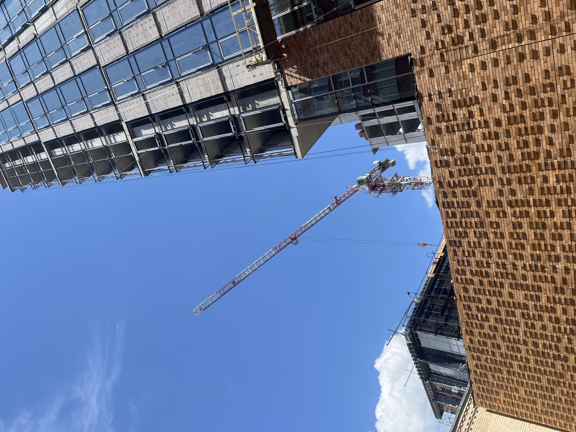 This is an image taken from a low angle, looking up at a bright blue sky with some wispy white clouds. The scene depicts a construction site with several large buildings and a construction crane.
In the foreground, on the right, is a building with a facade made of textured reddish-brown brick. The brickwork has a unique pattern of protruding blocks that cast distinct shadows. A section of this building has large, dark-tinted windows.
To the left of the brick building, and partially overlapping it, is another building under construction. This building features a facade of glass windows set within a concrete and metal frame. The windows reflect the sky and surrounding structures. The lower levels of this building show exposed metal scaffolding and concrete supports, indicating ongoing construction.
Dominating the sky is a large construction crane. Its boom is extended diagonally across the frame, from the upper right towards the lower left. The crane is red and white, with a lattice-like structure.
The overall impression is one of urban development and industrial activity. The bright sunlight suggests it is daytime, likely midday, given the intensity of the light and the shadows. The clear blue sky and scattered clouds indicate fair weather. While no people are directly visible, the presence of the construction crane and buildings implies human activity related to construction. There is no visible text in the image. The location context is Dnipro, Ukraine.