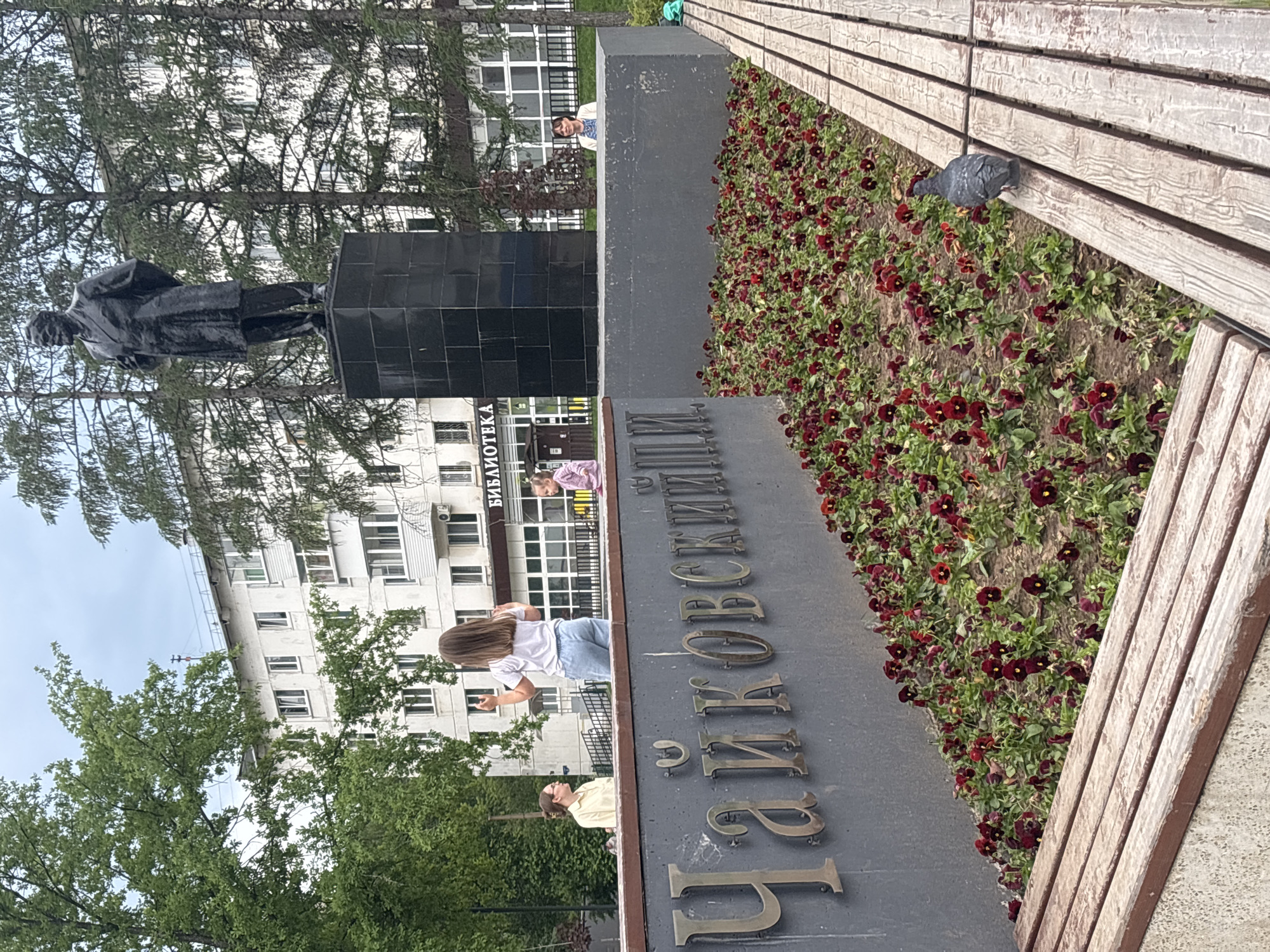 A pigeon stands on a wooden bench in front of a flower bed filled with dark red flowers. Behind the flowers is a gray wall with large metal letters spelling out "ЧАЙКОВСКИЙ". In the background, a statue of a man in a suit stands on a black pedestal. To the left of the statue is a white building with many windows, and trees with green leaves. A woman in a light-colored shirt and light blue jeans is standing in front of the wall, facing away from the camera and gesturing with her hands. Another woman in a yellow shirt is standing to her left, and a child in a pink outfit is standing to her right, near the statue. The sky is overcast.