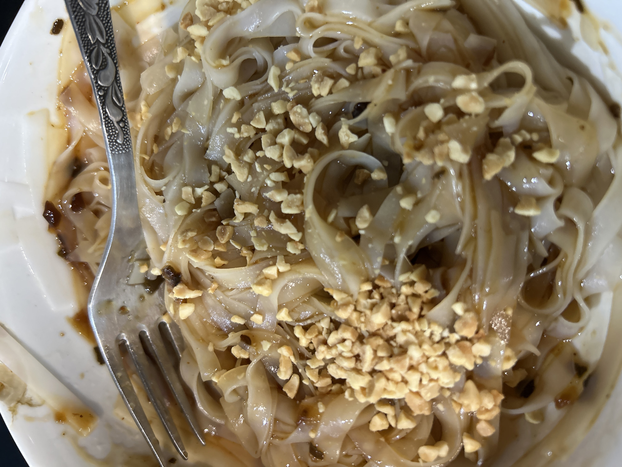 A close-up, overhead shot displays a dish of flat, wide noodles served in a white bowl, likely in Chaikovsky, Russia. The noodles are coated in a glossy, dark brown savory sauce, possibly peanut or soy-based, with visible specks of dark ingredients, possibly minced garlic or fried shallots. A generous amount of roughly chopped peanuts is sprinkled over the noodles, forming distinct clusters. A silver-colored fork with an ornate, leaf-patterned handle is positioned diagonally across the bowl, with its tines partially submerged in the noodles and sauce, suggesting the dish is being, or is about to be, eaten. The white bowl shows some residue of the sauce on its interior edges, indicating prior use or stirring. The lighting appears to be ambient indoor light. There is no visible text.