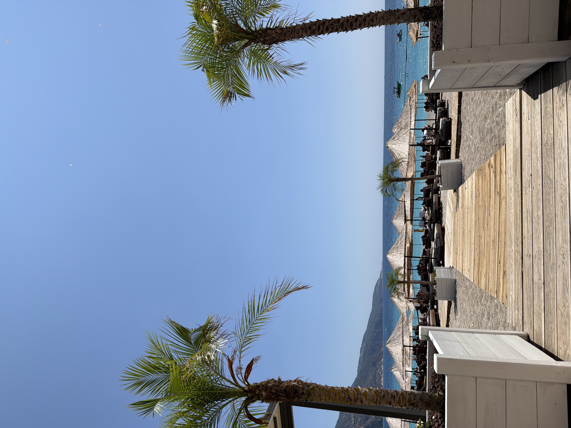 A tranquil beach scene in Fethiye, Turkey, is captured under a clear blue sky. Two prominent palm trees frame the view, one on the left and another on the right. A wooden pathway leads towards a stretch of beach lined with thatched umbrellas and lounge chairs, indicating a resort or recreational area. The turquoise water of the sea stretches to the horizon, where a faint outline of mountains can be seen. Small boats dot the water, and a few paragliders are visible in the sky above, suggesting a popular spot for both beach activities and adventure sports. The presence of planters and white wooden barriers suggests a well-maintained and organized beach area, likely catering to tourists seeking relaxation and leisure.