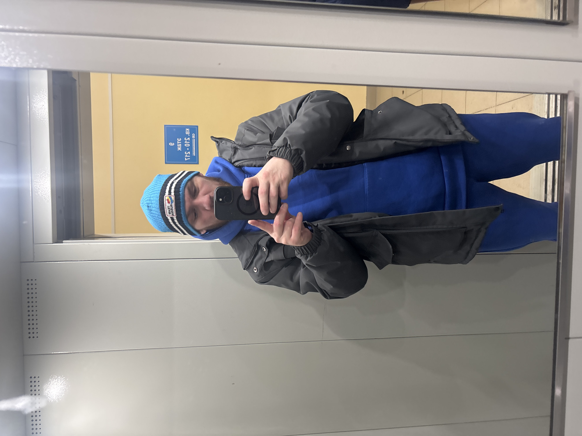 The image shows a man in an elevator, reflected in a mirror. He is wearing a blue beanie with white and black stripes, a blue hoodie, and a dark-colored jacket. He is holding a phone and taking a selfie. The elevator has a yellow wall with blue signage. The sign is written in Russian, "ЖИДС KB 310-SJ1 Э". This suggests the elevator is located in Perm, Russia. The interior of the elevator is generally gray.