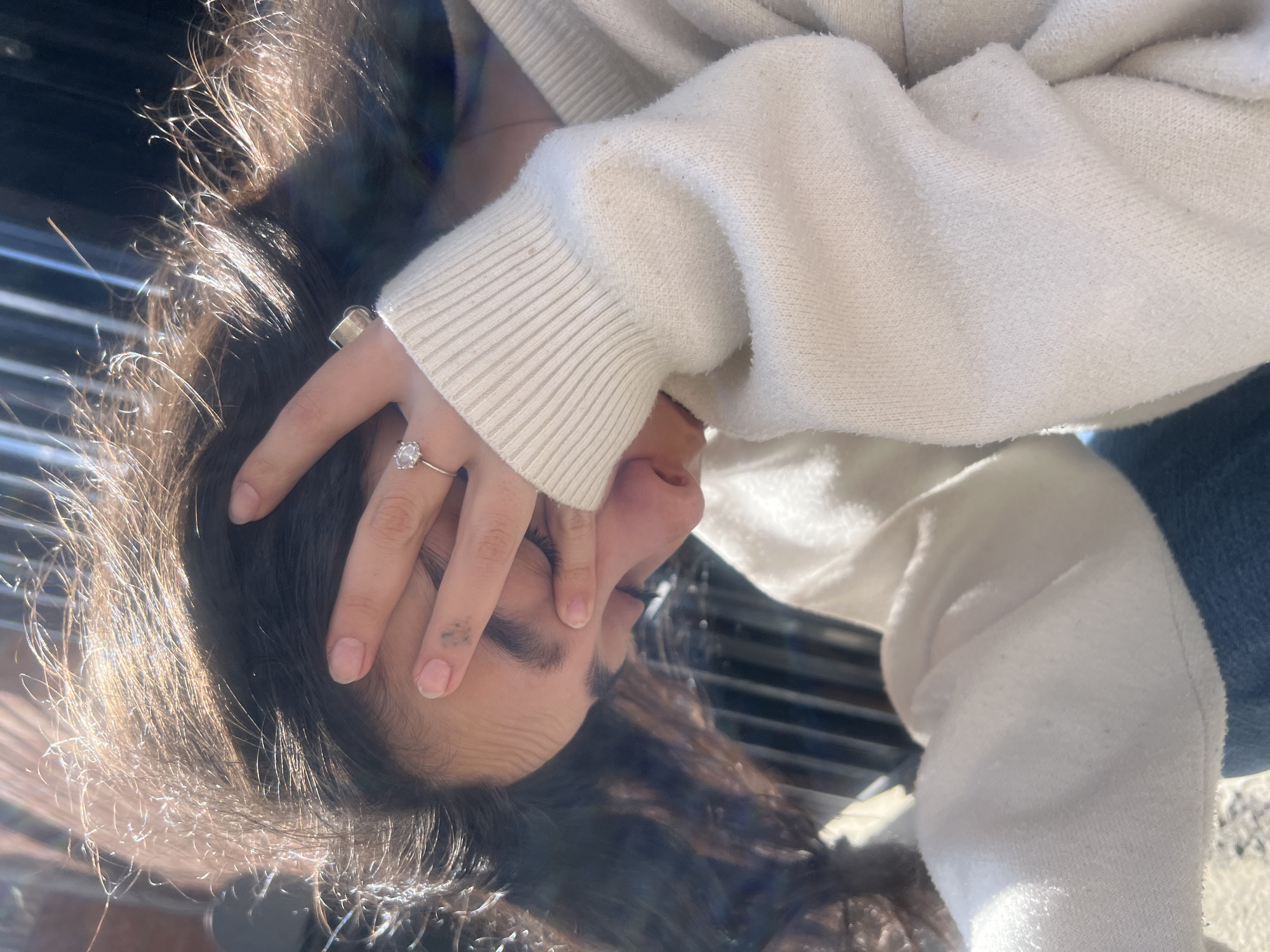 A person is outside in Tbilisi, Georgia. They are wearing a white hooded sweatshirt and denim pants. They are covering their face with their left hand. They are wearing two rings on their left hand. The lighting suggests the photo was taken during daylight hours. A structure with metal bars is behind the person. The ground is partially visible with a concrete like texture.