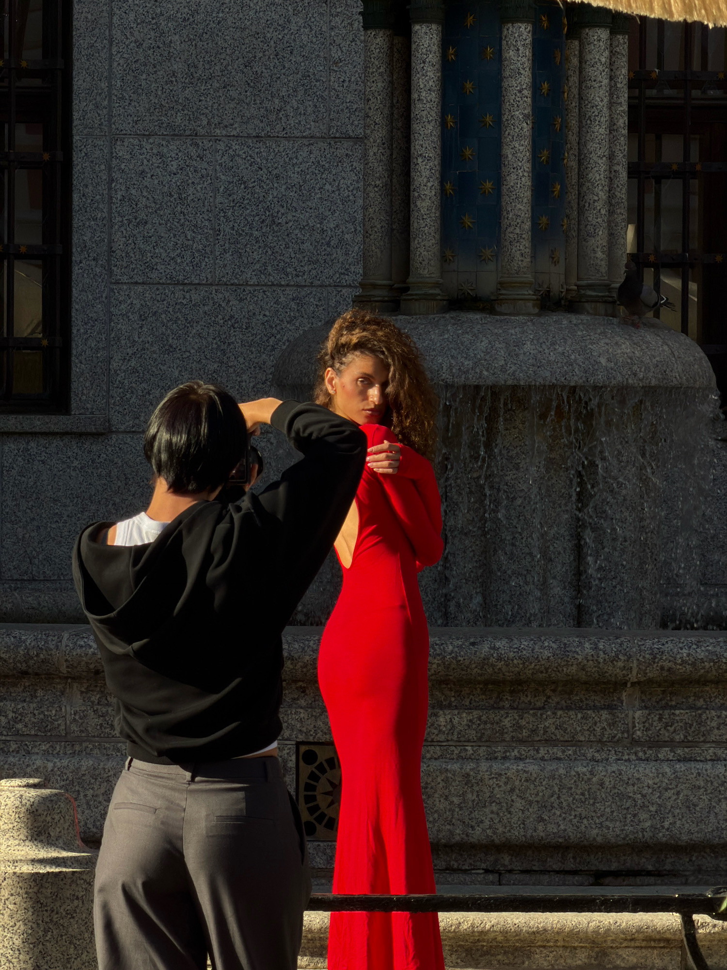 A person wearing a black hoodie and gray pants is taking a photograph of a woman in a long, bright red dress. The woman has curly brown hair and is looking towards the camera. She is standing in front of a fountain with water cascading down its stone structure. Behind the fountain are decorative columns with blue and gold star patterns. The background also includes a stone building with windows and a metal grate. The scene appears to be outdoors in a city.
