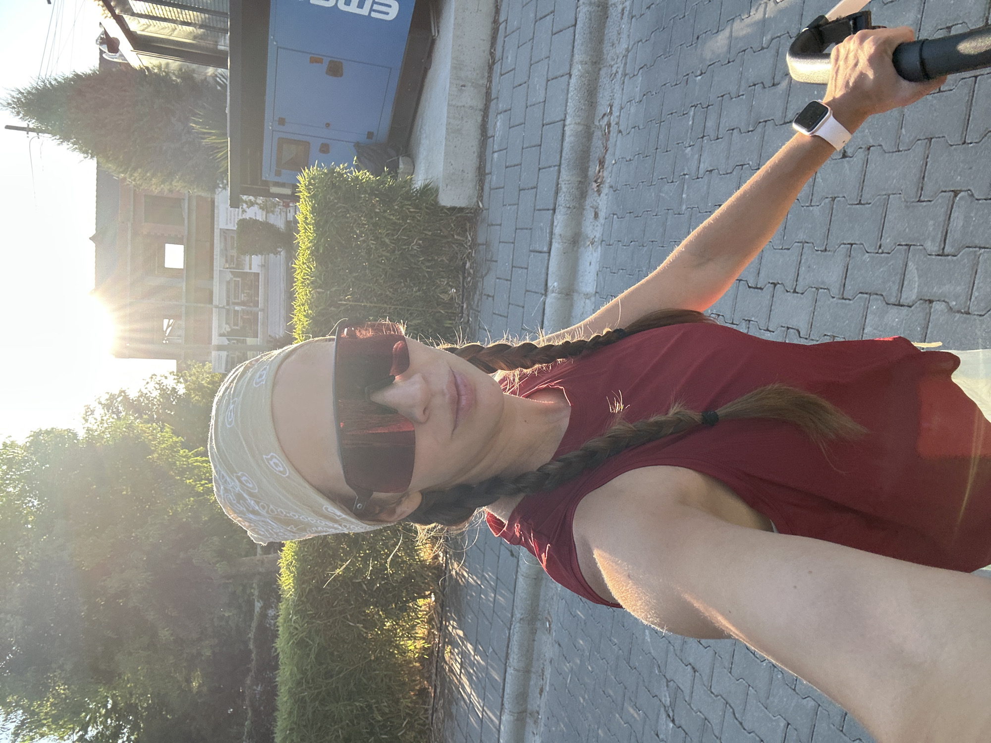 A woman is taking a selfie while outdoors, possibly in Fethiye, Turkey, during daylight. She is wearing a red tank top, a light-colored bandana on her head, and large sunglasses. Her hair is styled in two braids. In the background, there is lush green foliage, a building under construction, and a large blue generator. The ground is paved with interlocking bricks. The sun is shining brightly, casting shadows and creating lens flare in the image. The woman appears to be holding onto the handles of a stroller or similar device, suggesting she might be on the move.