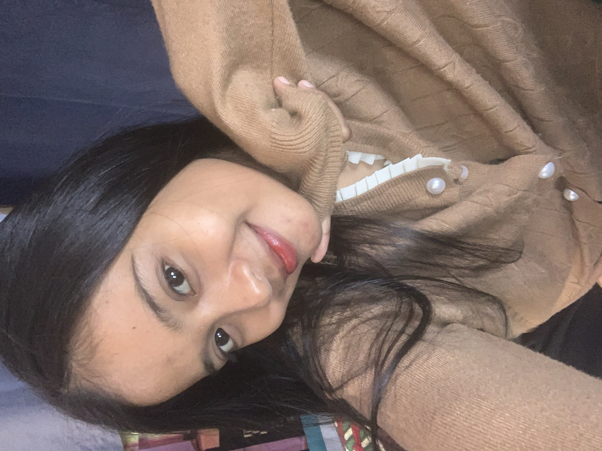 The image is a close-up selfie of a young woman with long, dark hair and brown eyes. She is wearing a light brown, cable-knit cardigan with pearl buttons and a white ruffled collar peeking out from underneath. Her right hand is resting on her chin, and she is smiling at the camera. The background is out of focus and appears to be an indoor setting with some colorful objects visible on the left side. The woman's skin has a warm tone, and she is wearing red lipstick. The lighting suggests an indoor environment, possibly with natural light. There is no discernible information in the image that can be used to determine its location in Sungai Meriam, Indonesia.