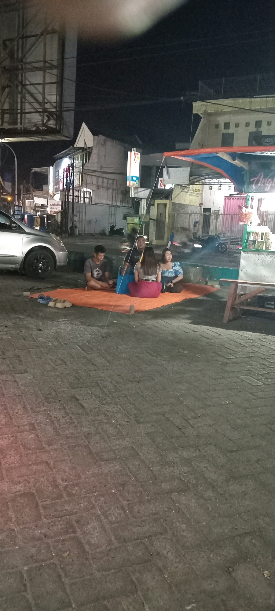The image depicts an outdoor scene at night in Babat, Indonesia. Four individuals are seated on an orange tarp spread on a cobblestone ground. One male, wearing a grey t-shirt, is on the left, facing forward. To his right are two females, one in a dark top and pink pants, and another in a blue floral top. Another male, wearing a dark shirt, is behind the first male, and appears to be facing towards the right. A silver car is parked to the left of the group. Behind them, there are buildings and streetlights, suggesting an urban environment. A canopy is visible overhead, supported by poles. The overall impression is of a casual gathering, possibly at a street-side food stall or resting area.