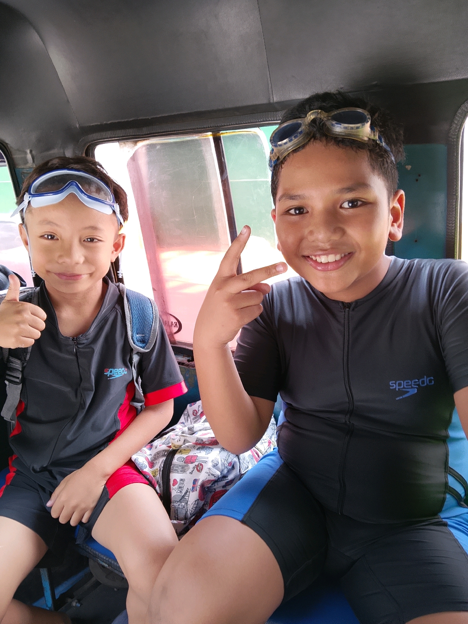 Two young boys, likely pre-teens, are seated in the back of a vehicle, possibly a type of open-air transport common in Southeast Asia. They are both wearing dark gray and blue swimming attire with goggles perched on their heads. The boy on the left is giving a thumbs-up, while the boy on the right is making a peace sign and smiling broadly. A backpack with a colorful, cartoon-like print is visible between them. The setting appears to be a bright, possibly sunny day, given the lighting and the overall cheerful mood. The vehicle's interior is visible, with dark gray paneling and windows. The image suggests they are on their way to or from a swimming activity, perhaps at a beach or pool, given their attire. The scene evokes a sense of fun and anticipation. There is visible text on the clothing of both boys: "Speedo" is clearly legible on the shirt of the boy on the right and a similar logo is on the shirt of the boy on the left. The text "MALANG" is visible on a pink label behind the boy on the left, indicating the location.