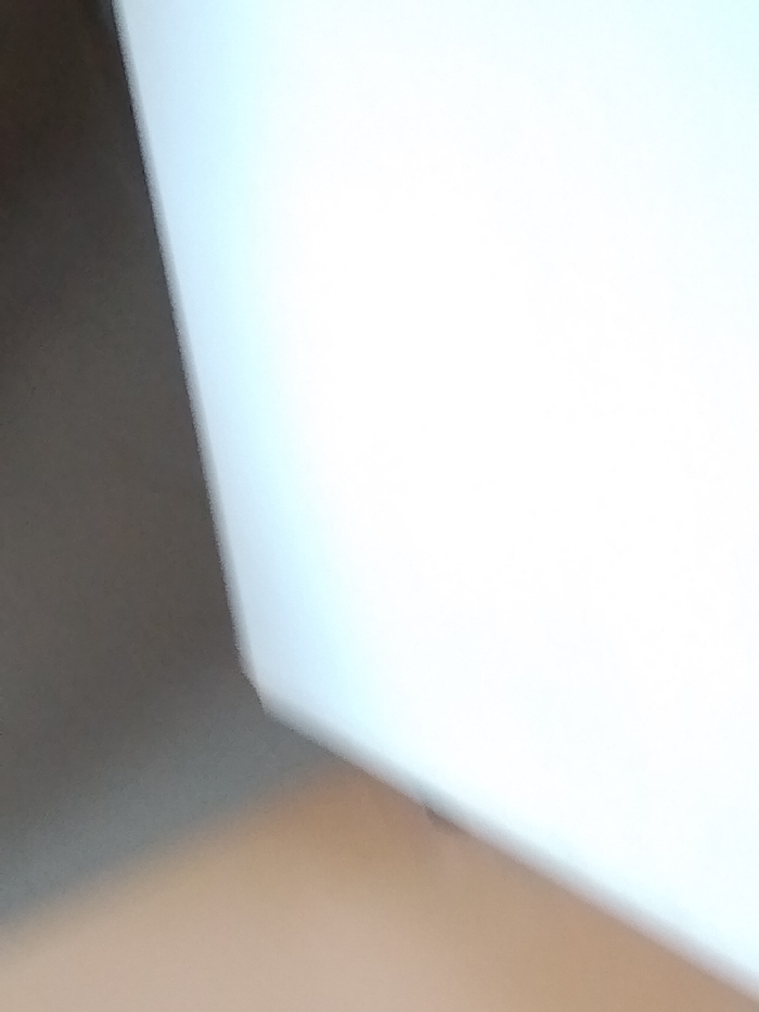 The image shows a close-up view of an indoor corner. The lower portion features a light brown surface, possibly a floor. A light gray wall extends diagonally upwards from the left. To the right of the wall is a white surface, probably another wall or ceiling. The scene is blurry. The location is unknown.