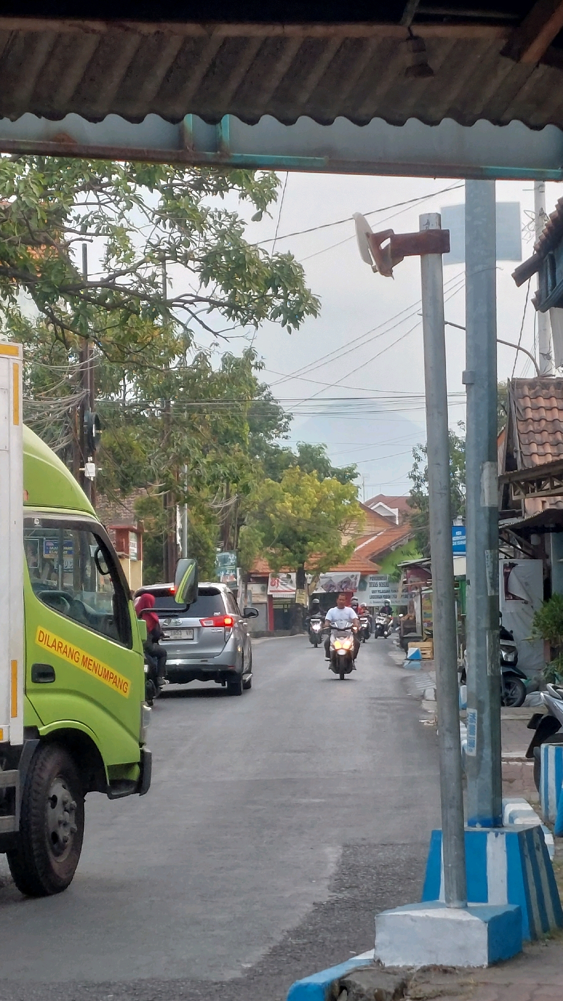 The image captures a street scene in Sidoarjo, Indonesia, likely during the day. In the foreground on the left, a green truck is partially visible, with yellow text on its side reading "DILARANG MENUMPANG". The truck appears to be in motion or stopped in traffic. Ahead of the truck, a silver SUV is in the lane, with a person wearing a red headscarf seated on the back. Further down the road, several motorcycles are traveling in the same direction. The road is paved and appears to be a two-lane street lined with buildings on the right, featuring tiled roofs characteristic of Indonesian architecture. Trees with green foliage are present on the left and in the background. A utility pole with wires is prominent on the right side of the road, along with concrete barriers painted with blue and white stripes. In the far background, a hazy mountain or hill is visible. The overall impression is a typical urban or semi-urban street environment in Indonesia with moderate traffic.