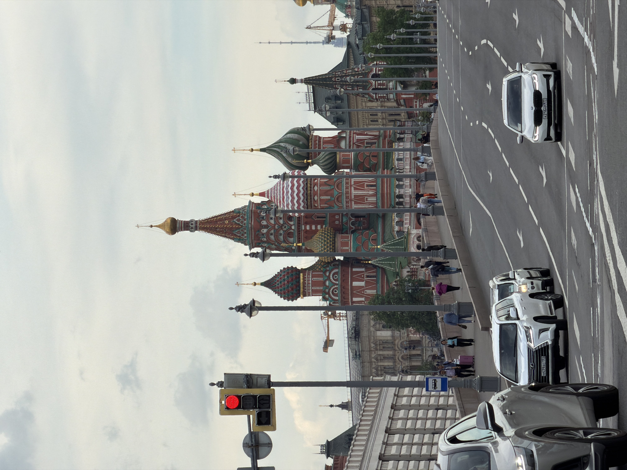 The image depicts a street scene in Moscow, Russia, with the iconic St. Basil's Cathedral in the background. A street with cars traveling on it runs parallel to the sidewalk where pedestrians are walking. A traffic light is visible in the foreground, currently displaying a red light. Several cars of different colors are stopped at the traffic light. The sky is overcast.