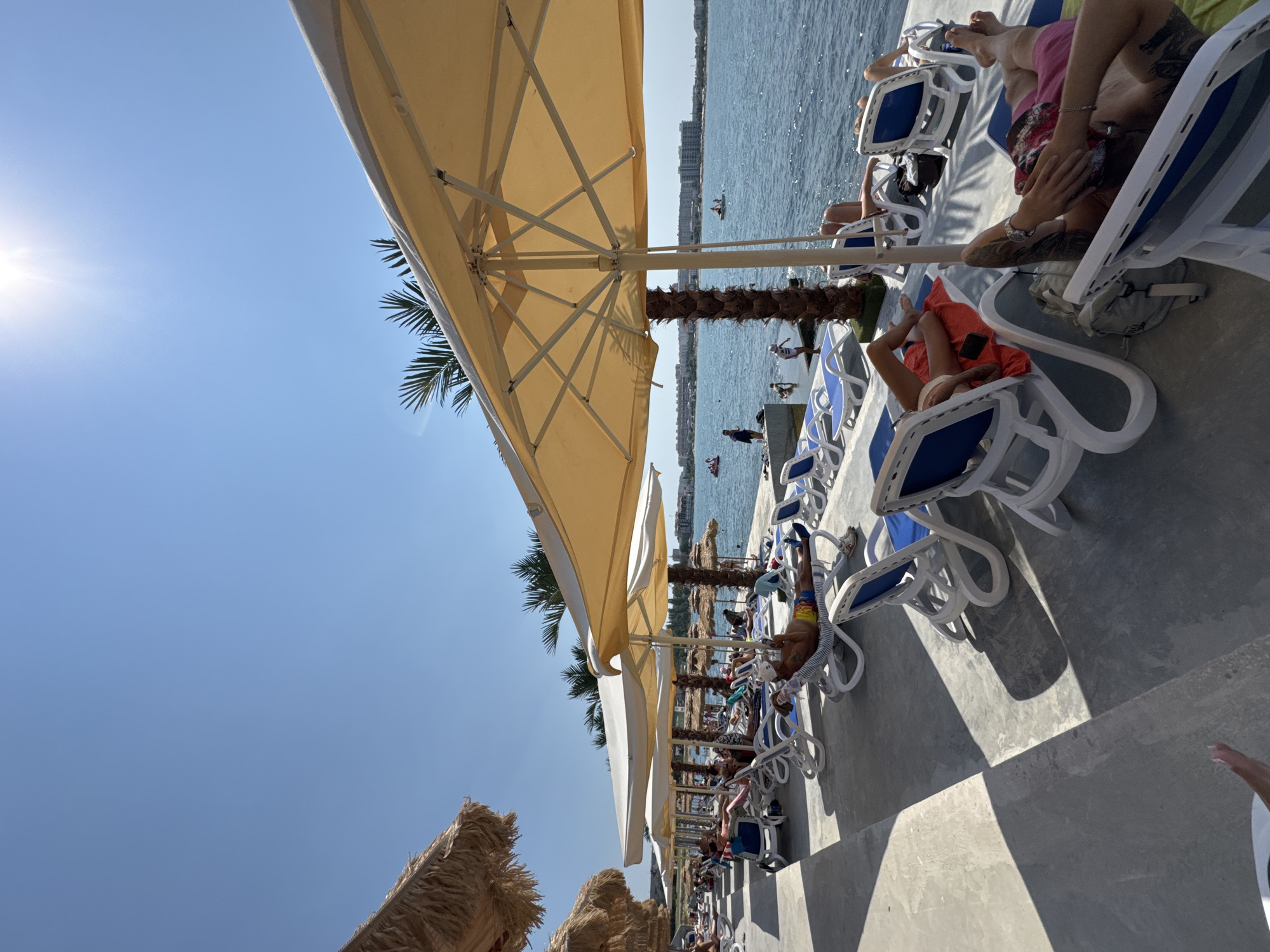 The image depicts a sunny day at a beach resort in Sevastopol, Ukraine. A row of white lounge chairs with blue cushions are arranged on a concrete patio, some occupied by people sunbathing. Large, light-colored umbrellas provide shade. Palm trees are visible behind the lounge chairs. In the background, the blue sea stretches towards a distant coastline with buildings. Several people are visible in the water, including some on inflatable toys and a person on a jet ski. The overall scene is one of leisure and relaxation in a warm, coastal environment.
