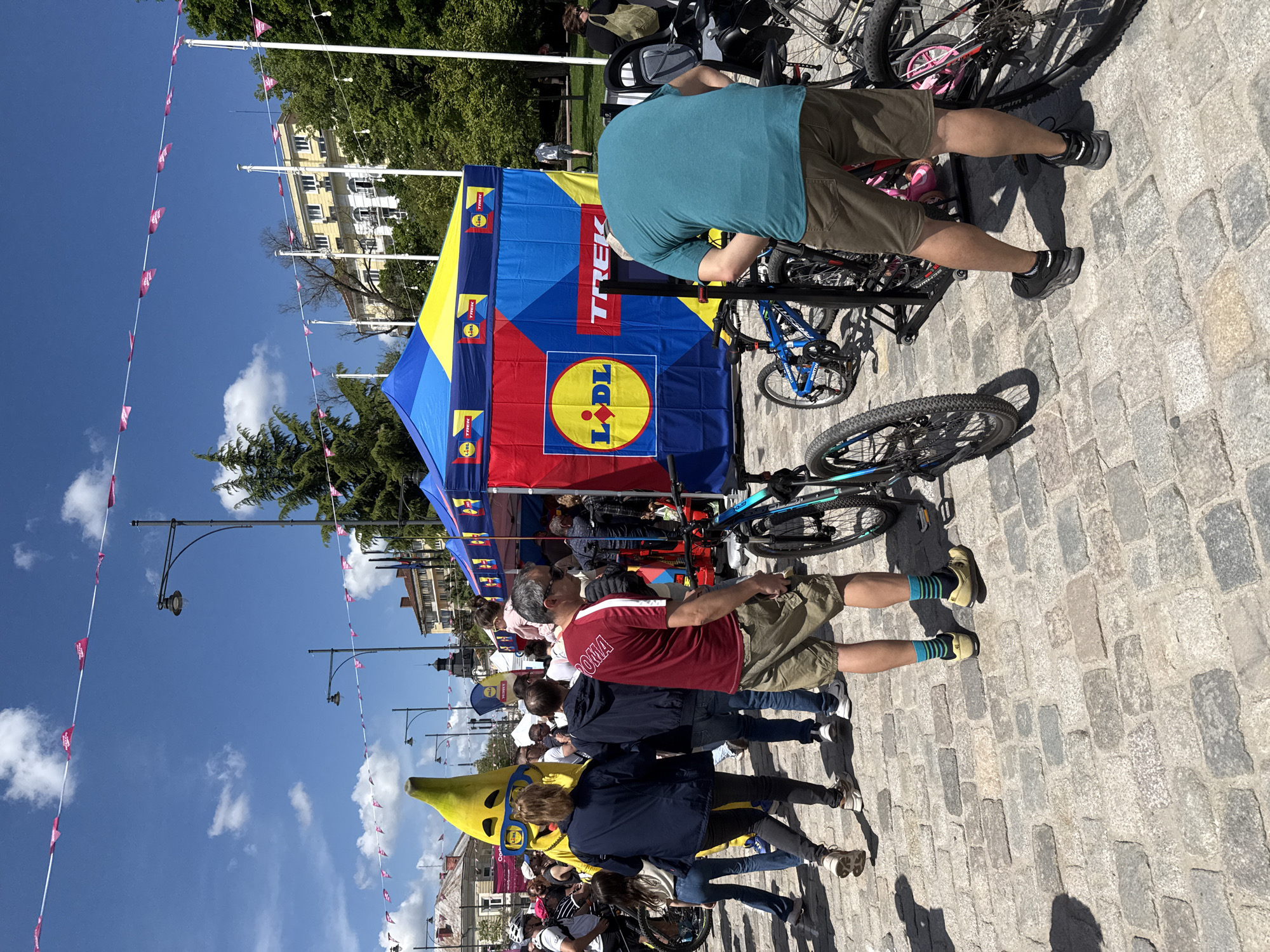 The image shows a street scene in Sofia, Bulgaria. The ground is paved with cobblestones. A crowd of people stands near a tent that displays the logos of Lidl and Trek. Bicycles are parked around the tent. Overhead, strings of pink flags are strung between lampposts. In the foreground, a person is dressed in a banana costume. Buildings and trees are visible in the background. The sky is blue with scattered clouds.