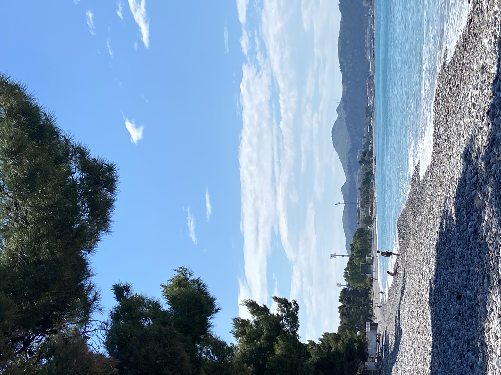 The scene depicts a sunny day at a pebble beach in Bar, Montenegro. Turquoise sea water laps against the shore, where several people are scattered. Two people are standing near the water's edge, while others are further down the beach, some appearing to sunbathe. Trees frame the left side of the view, partially obscuring the sky. In the background, mountains rise above a town and infrastructure that suggests a cityscape. The sky is partly covered with wispy clouds.