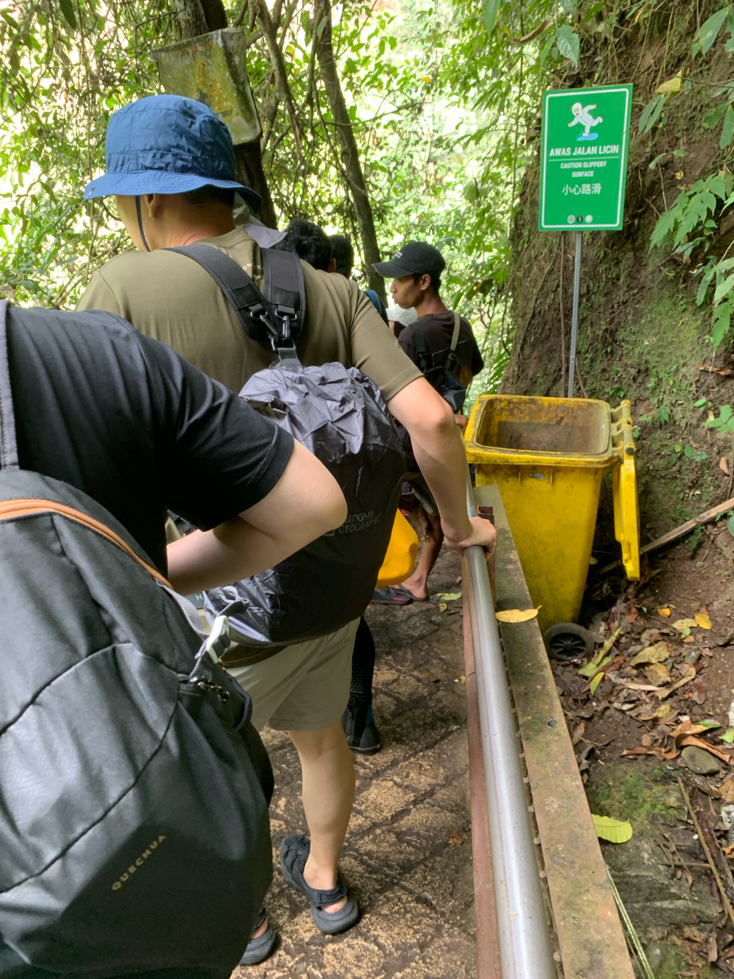 In the city of Sidomulyo, Indonesia, on a pathway surrounded by lush vegetation, a group of individuals are seen walking. One person in the foreground, partially obscured, wears a black t-shirt and carries a backpack. To their right, another individual in a green t-shirt and a blue bucket hat also carries a backpack. Behind them, another person in a black baseball cap walks, carrying a bag. A prominent yellow trash can is situated on the right side of the path, near a green sign displaying cautionary text in multiple languages, including "AWAS JALAN LICIN," "CAUTION SLIPPERY SURFACE," and "小心路滑." The pathway appears to be made of stone or concrete, with a metal railing on the right. The overall environment suggests an outdoor recreational area or a hiking trail.