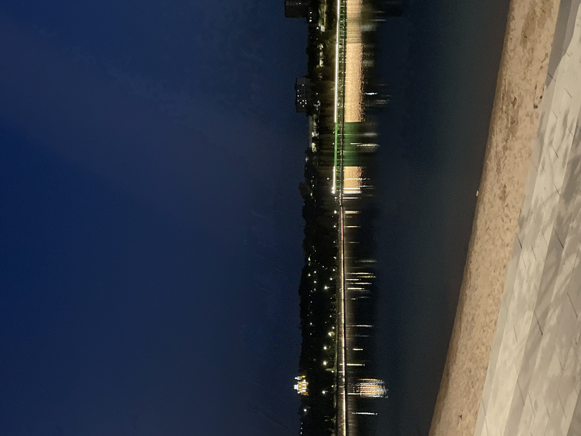 This image captures a nighttime scene at Lake Sunkar in Almaty, Kazakhstan. The dark blue sky is visible at the top, with faint light streaks possibly indicating rain or a lens artifact. Below the sky is a dark body of water reflecting the lights from the city on the opposite shore. The shoreline features a sandy beach and a paved walkway in the foreground. Along the distant shore, illuminated buildings and a long structure with green and white lights are visible. The lights create bright reflections on the water, with one particularly strong reflection of a building on the left. The overall atmosphere is serene and nocturnal, highlighting the city lights against the dark natural environment.