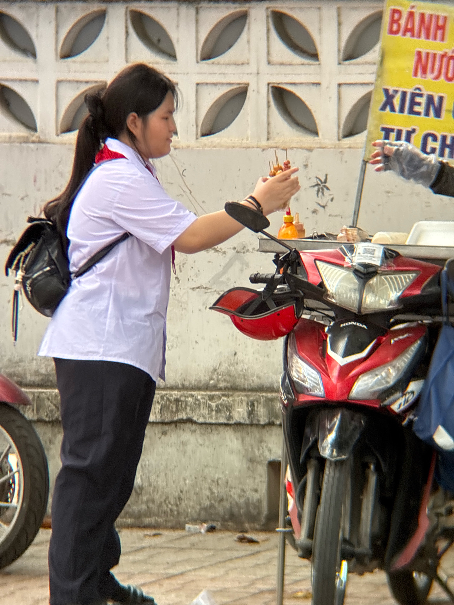 In Nha Trang, Vietnam, a young Vietnamese female, wearing a white short-sleeved blouse, dark pants, and a red scarf, stands near a food cart. She is seen from the torso down, with a black backpack slung over her left shoulder. Her right hand is extended, holding a skewer of what appear to be small sausages or meatballs, and she is interacting with another person whose gloved hand is visible reaching towards the food. A red helmet is hanging from a hook on the side of a red and black scooter, which is parked beside her. Behind the scooter, a yellow food sign displays Vietnamese text. The scene takes place outdoors, with a textured, light-colored wall in the background and a concrete ground.